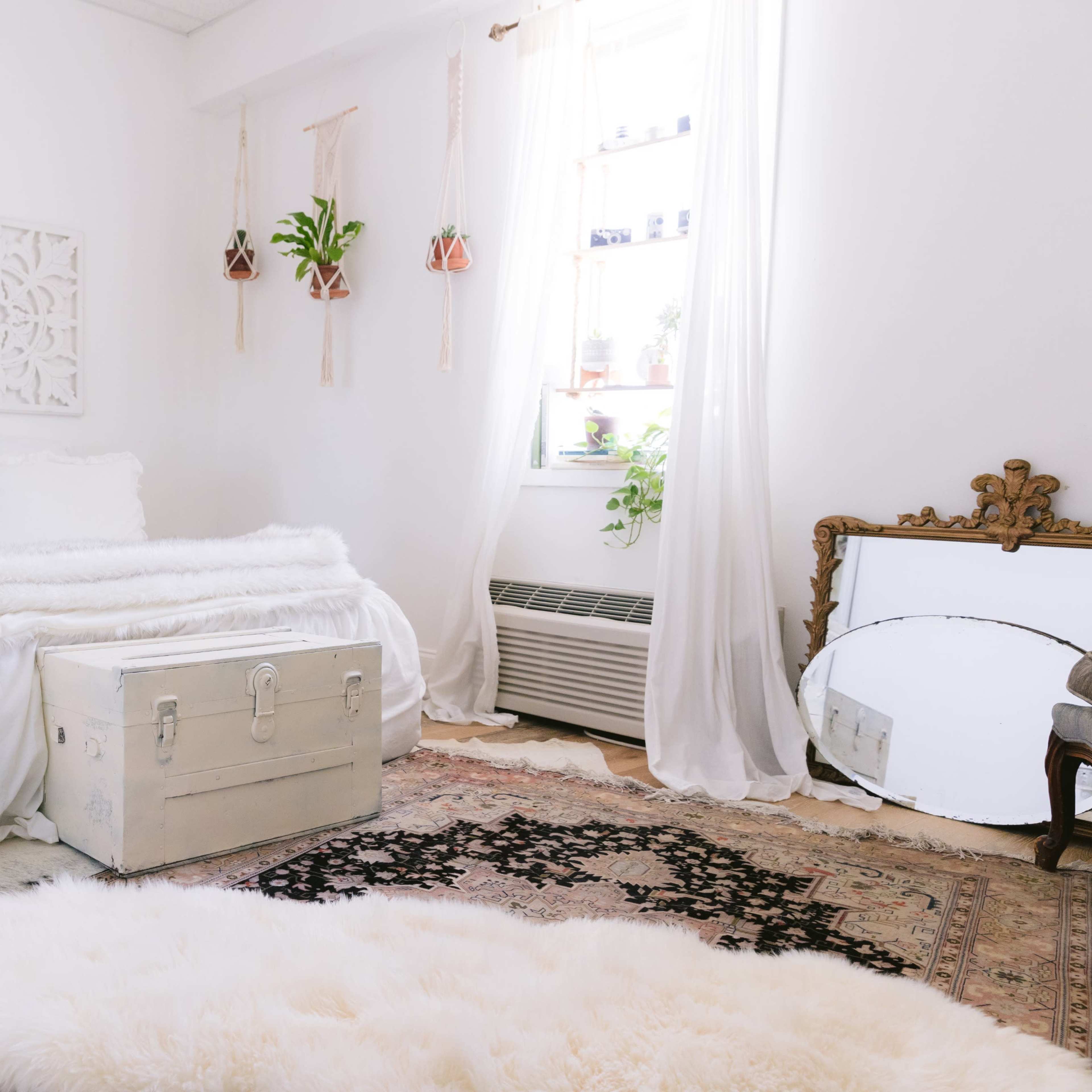 A bright and airy bedroom with a white bed covered in a fluffy blanket, a decorative wooden trunk at the foot, and plants on the windowsill.