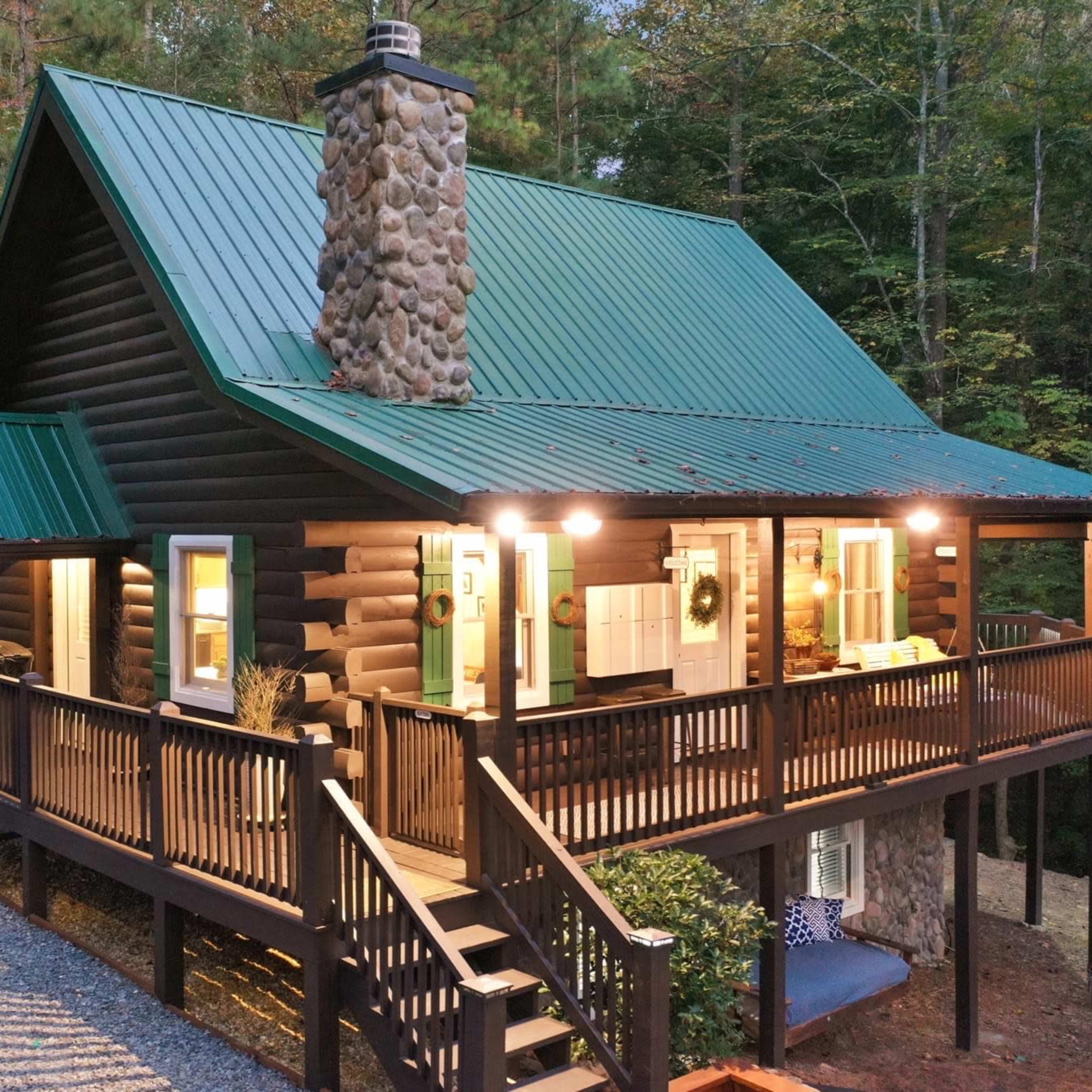 A log cabin with a green metal roof features a stone chimney and a spacious porch surrounded by trees.