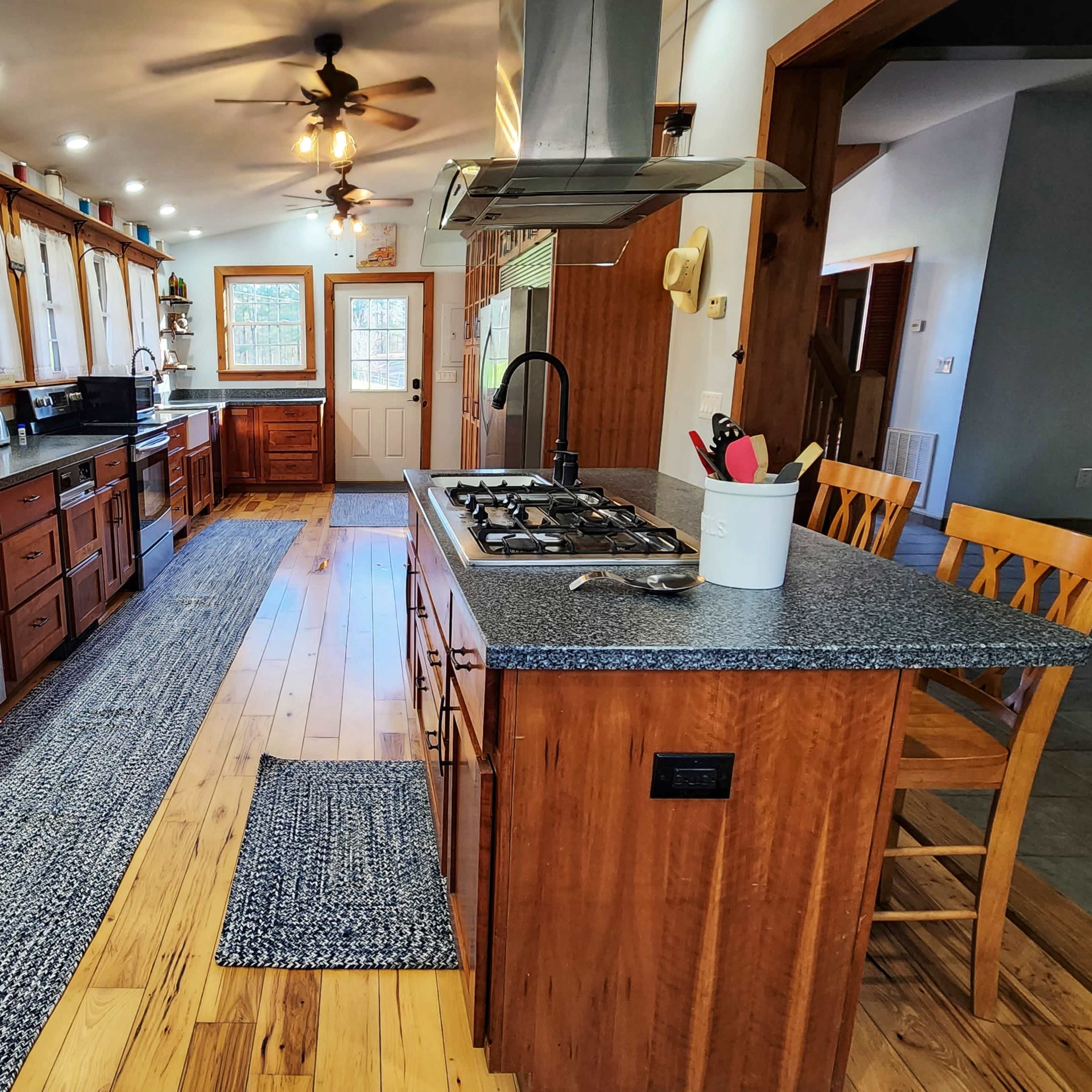 A modern kitchen with wooden cabinets, a central island featuring a gas stove, and large windows allowing natural light to enter the space.