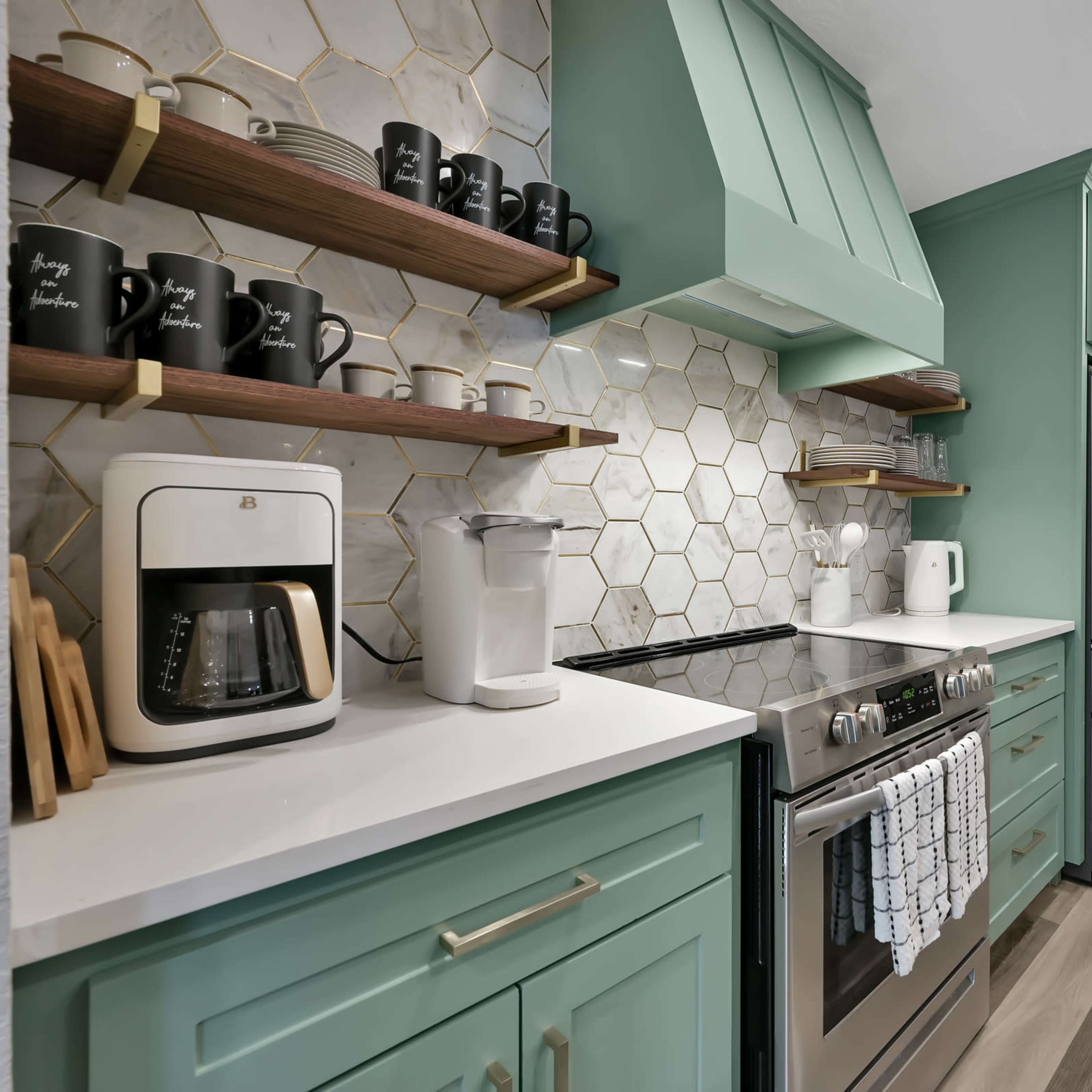 A modern kitchen features mint green cabinets, hexagonal tile backsplash, stainless steel appliances, and floating shelves with black mugs.