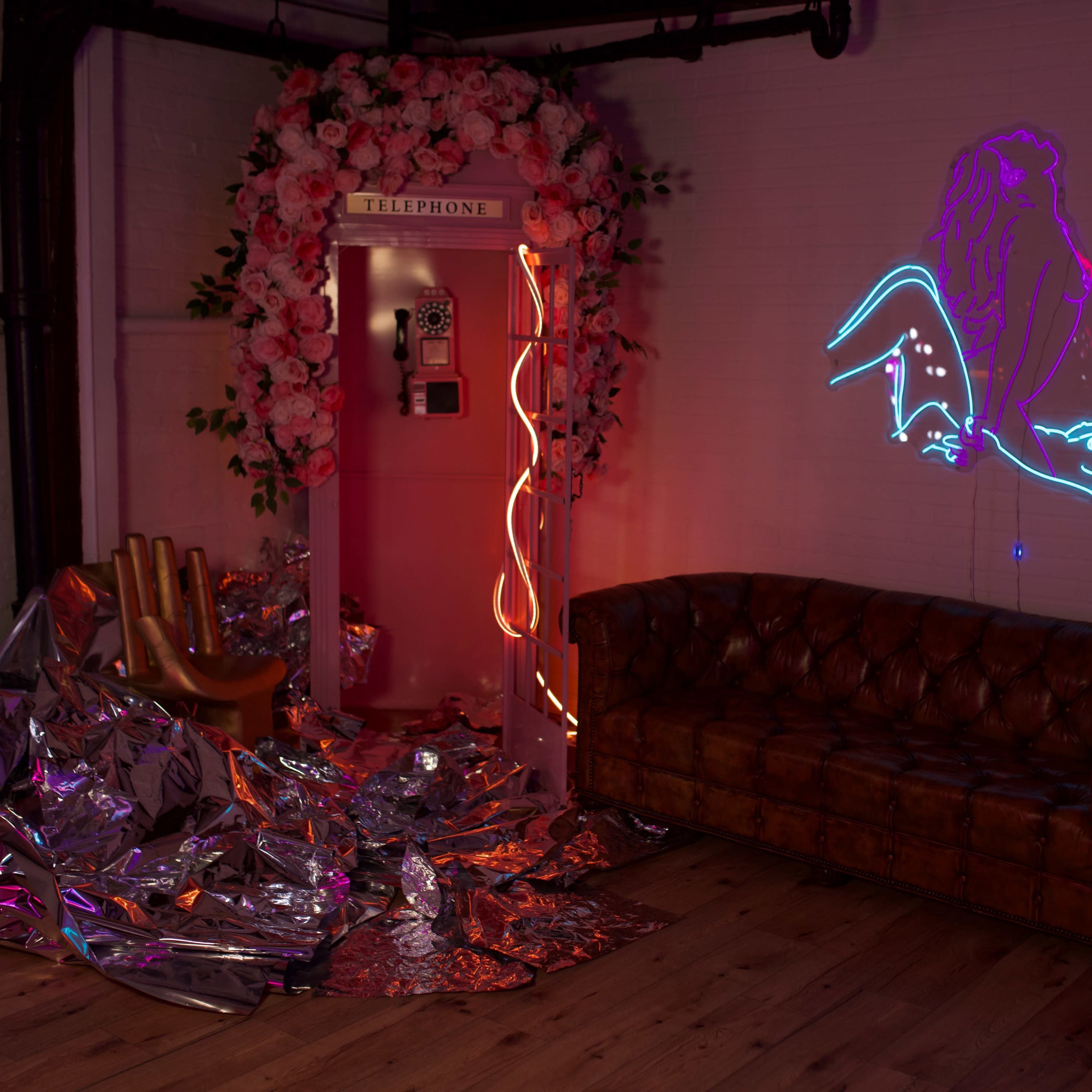 A vintage telephone booth surrounded by pink flowers, a neon lion graphic on the wall, a shiny silver material spread across the floor, and a brown leather sofa.