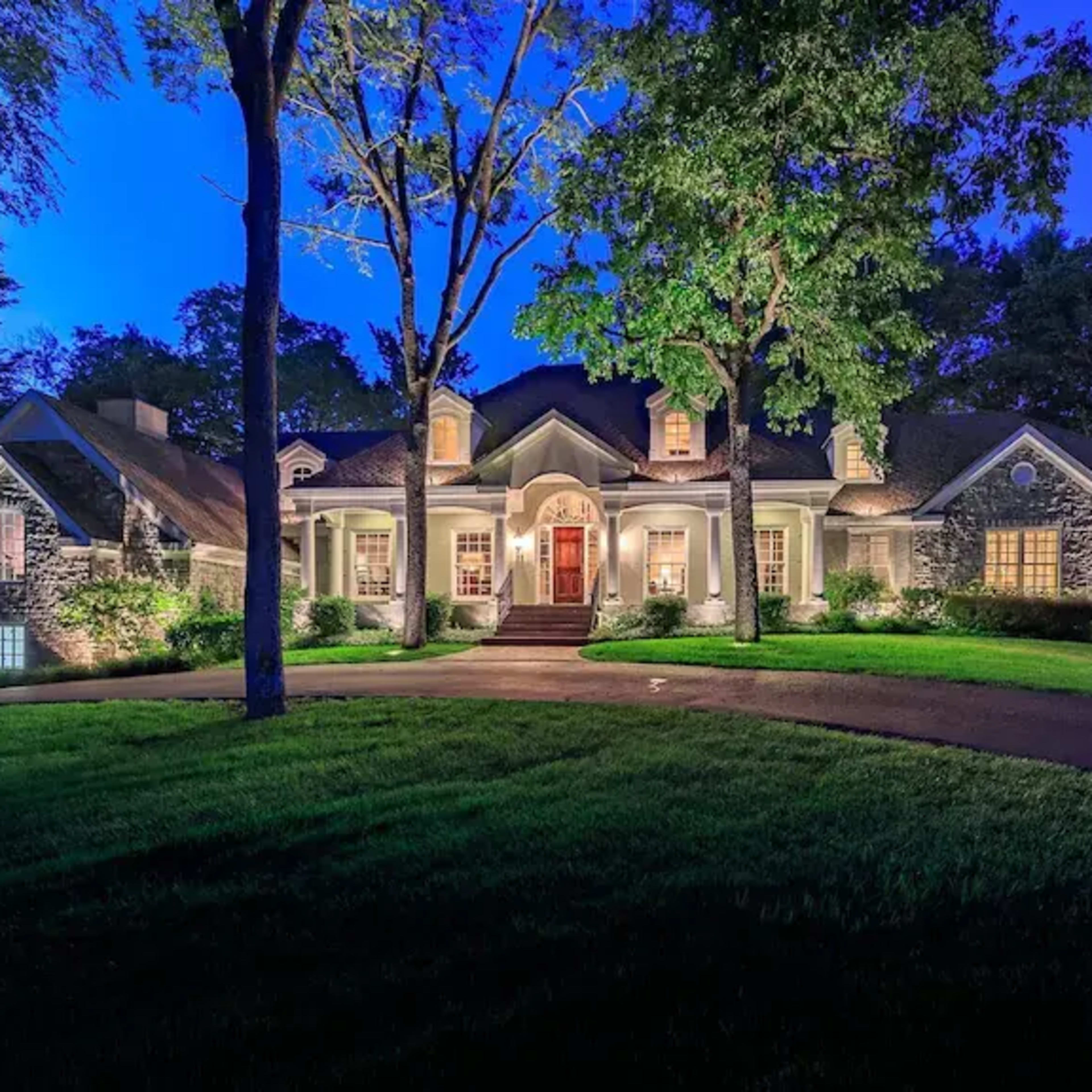 A large house with stone accents is illuminated at night, surrounded by trees and a well-manicured lawn.