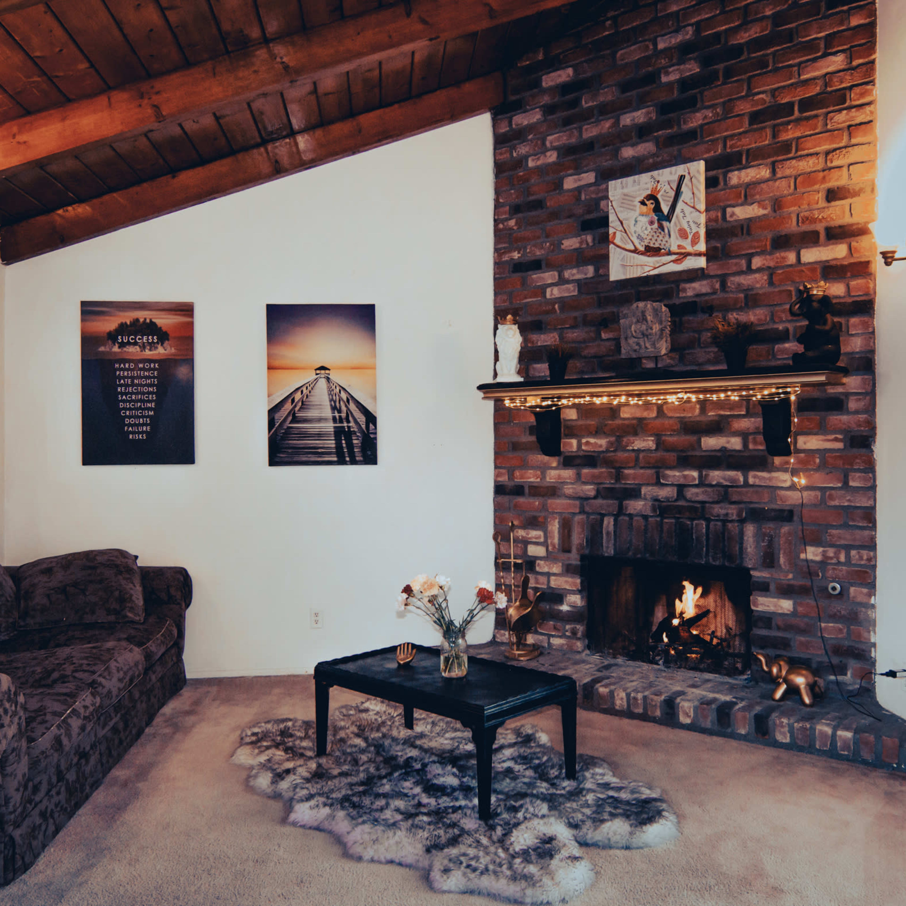 A cozy living room features a brick fireplace, a dark sofa, and two framed pictures on the walls, with a coffee table and a plush rug in front.