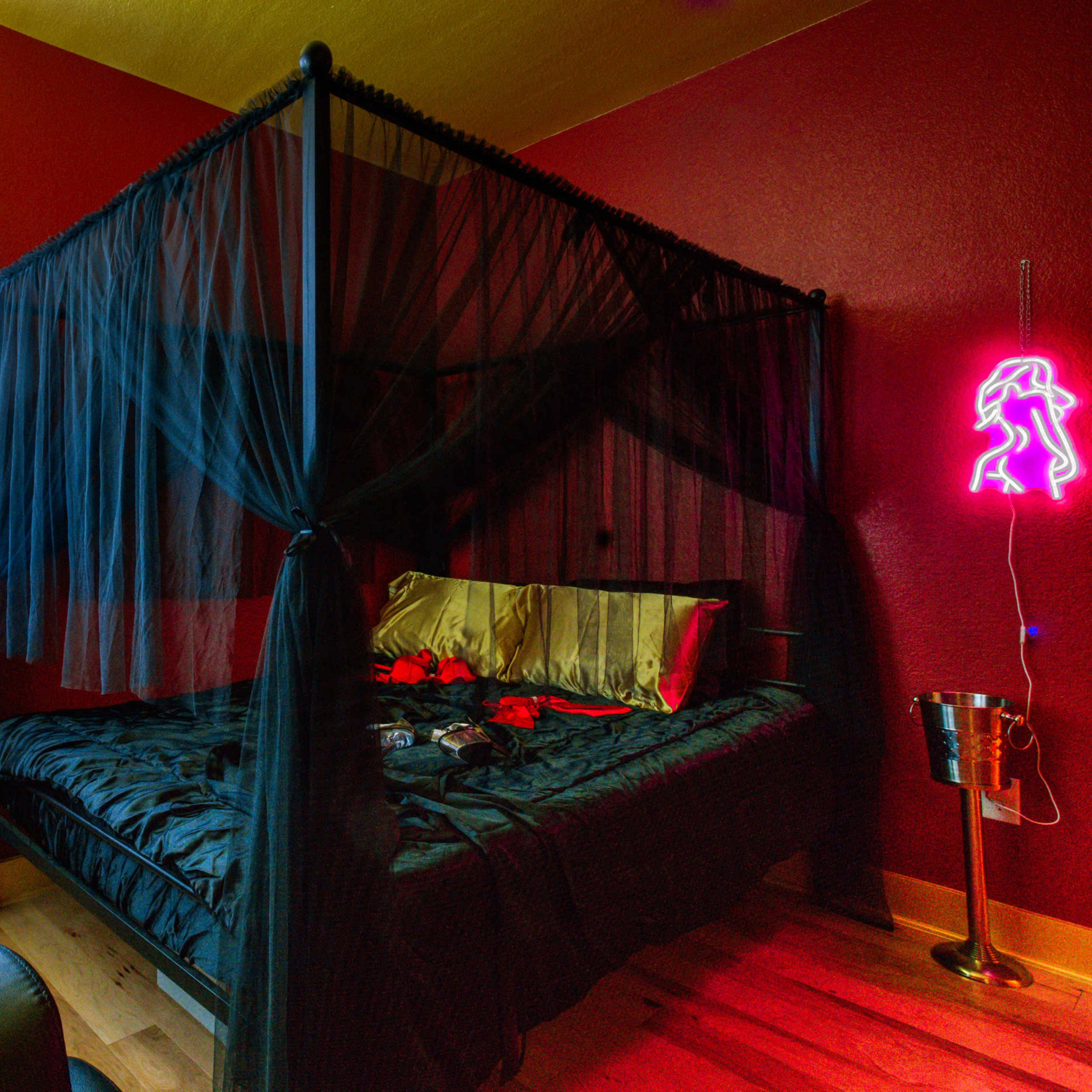 A canopy bed with sheer black drapes is positioned in a room with deep red walls, accompanied by a neon sign and a metal bucket.