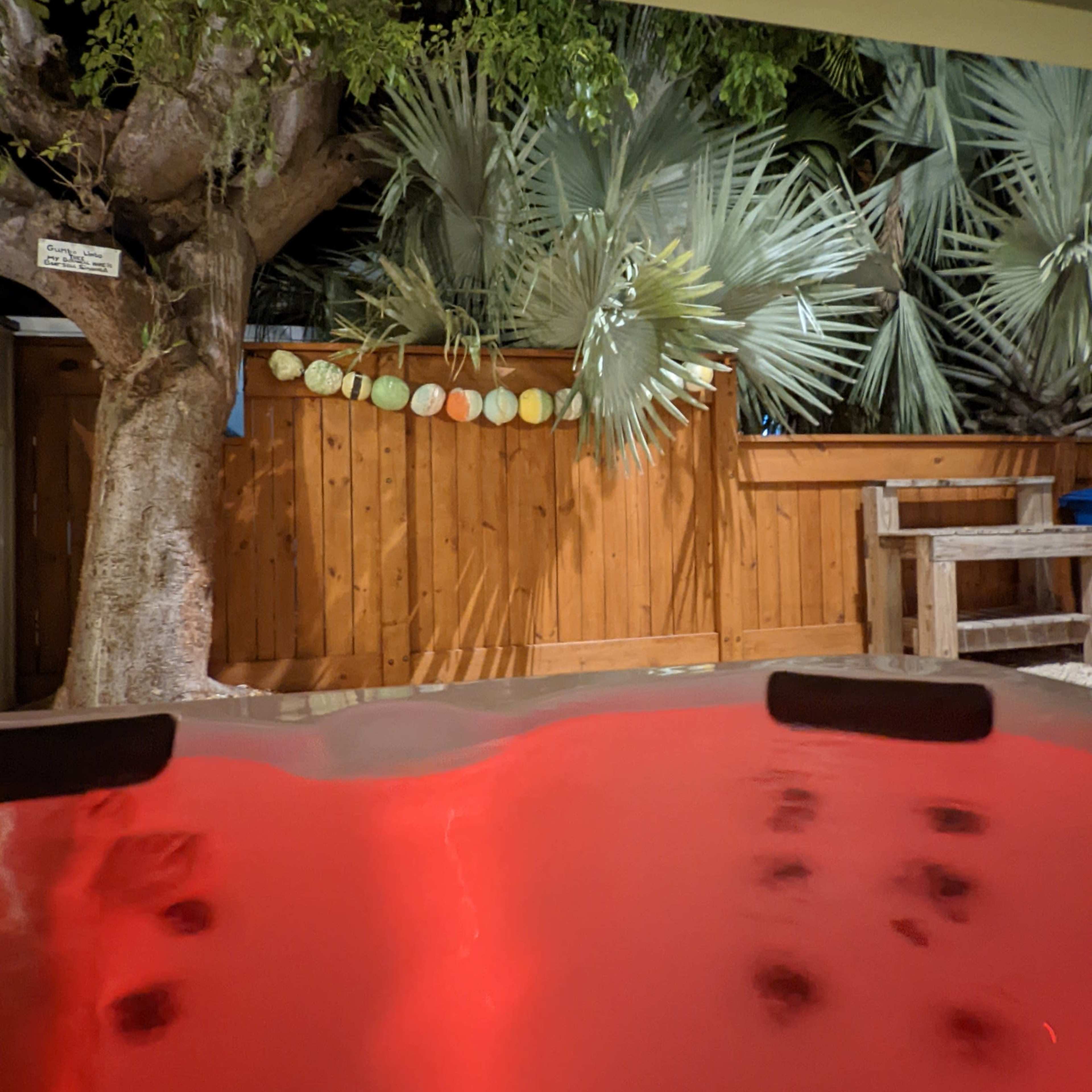 A hot tub with red lighting in the foreground, and a wooden fence with palm leaves and decorations in the background.