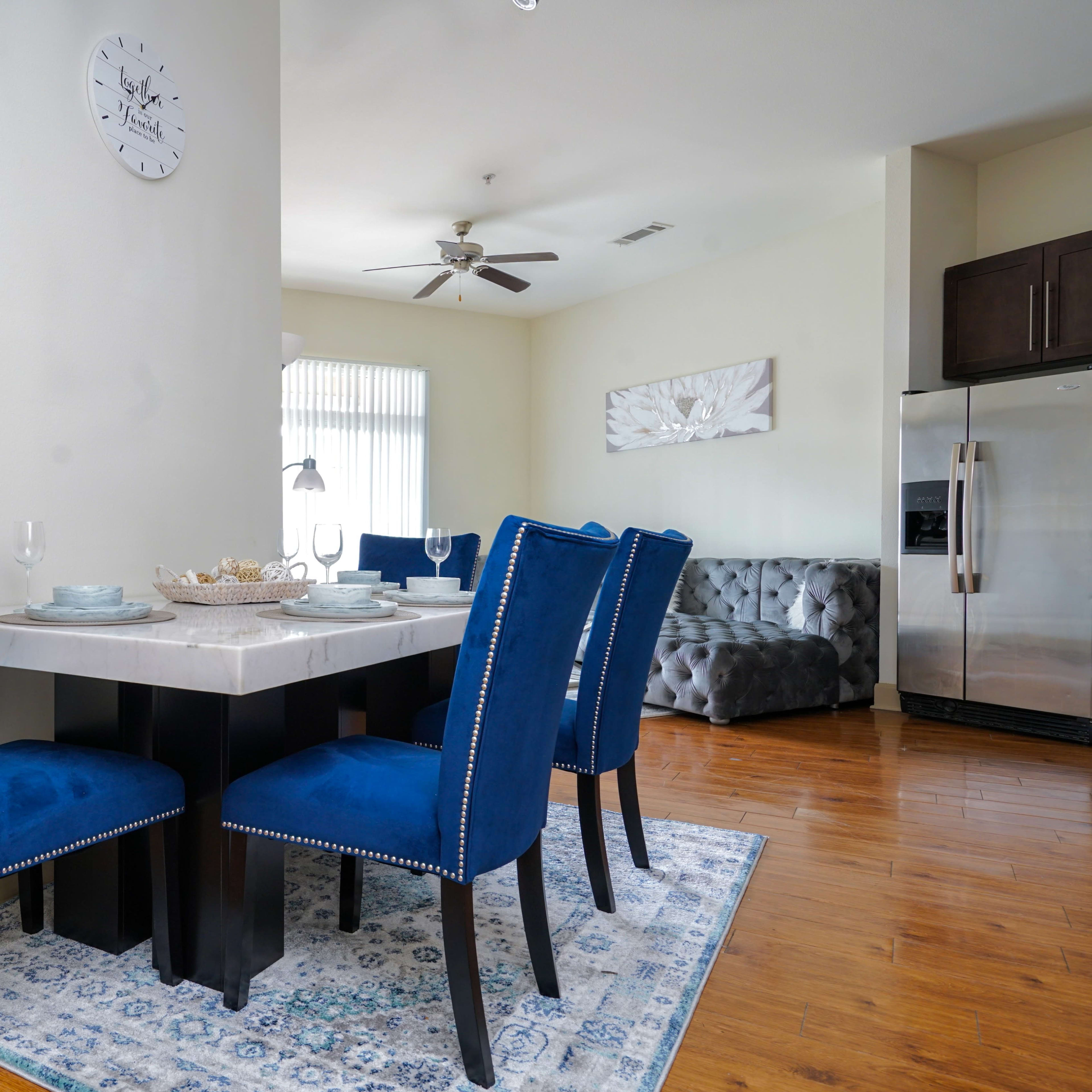A modern dining area features a marble-topped table surrounded by blue upholstered chairs, with a kitchen counter and a gray sofa visible in the background.