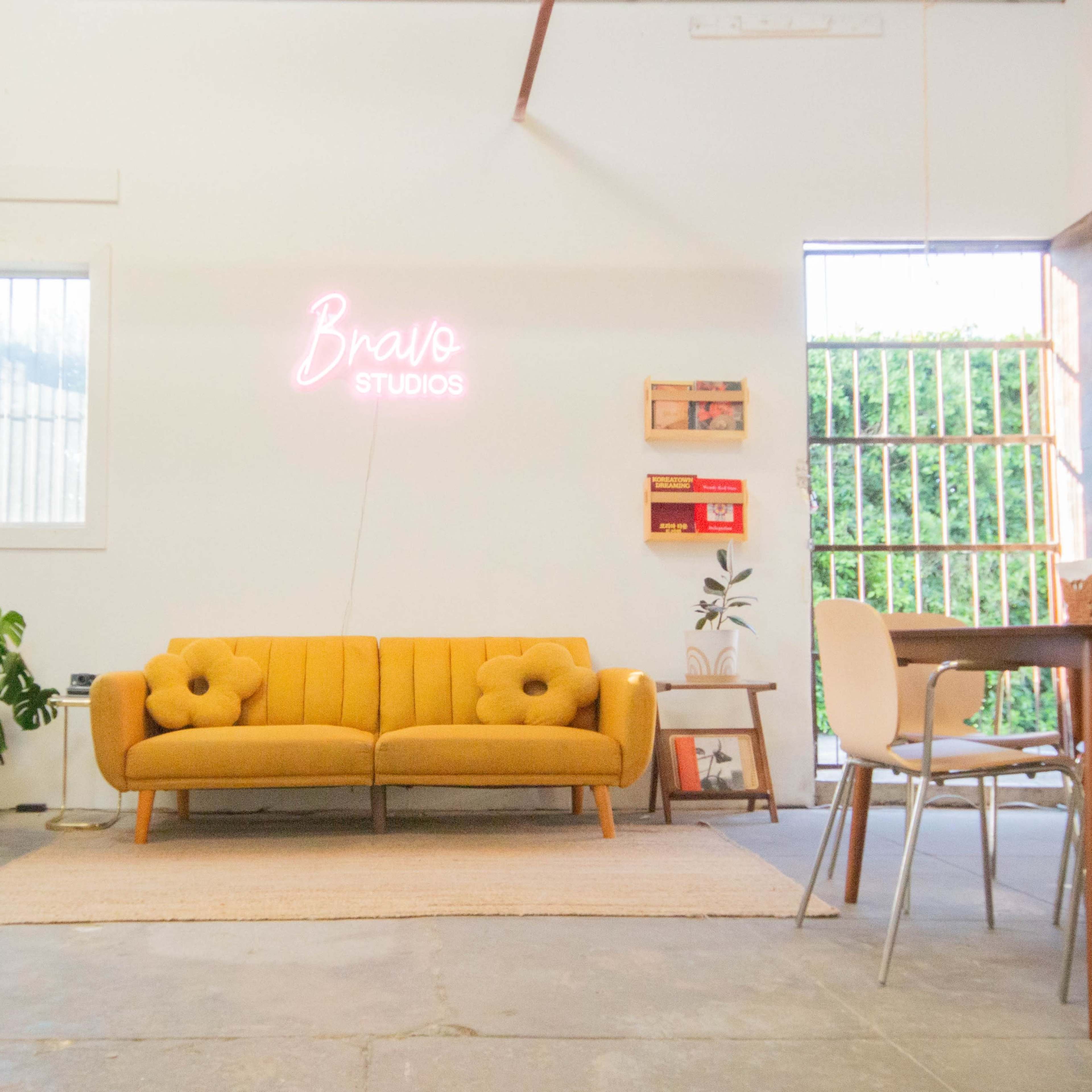A bright interior features a yellow couch, a dining table, and a neon sign that says "Bravo Studios."
