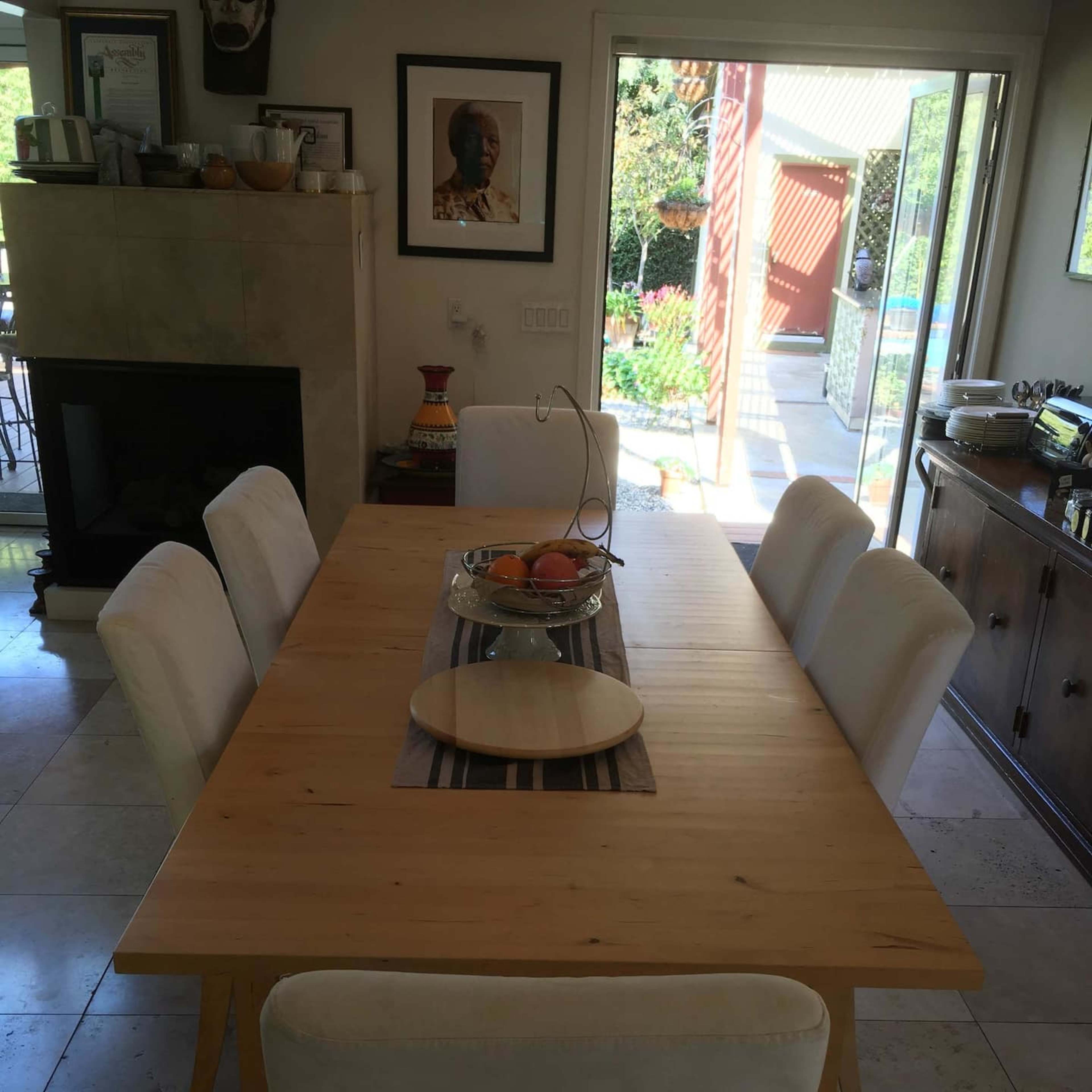 A wooden dining table with six upholstered chairs is set in a room with a fireplace, leading to a patio with outdoor views.