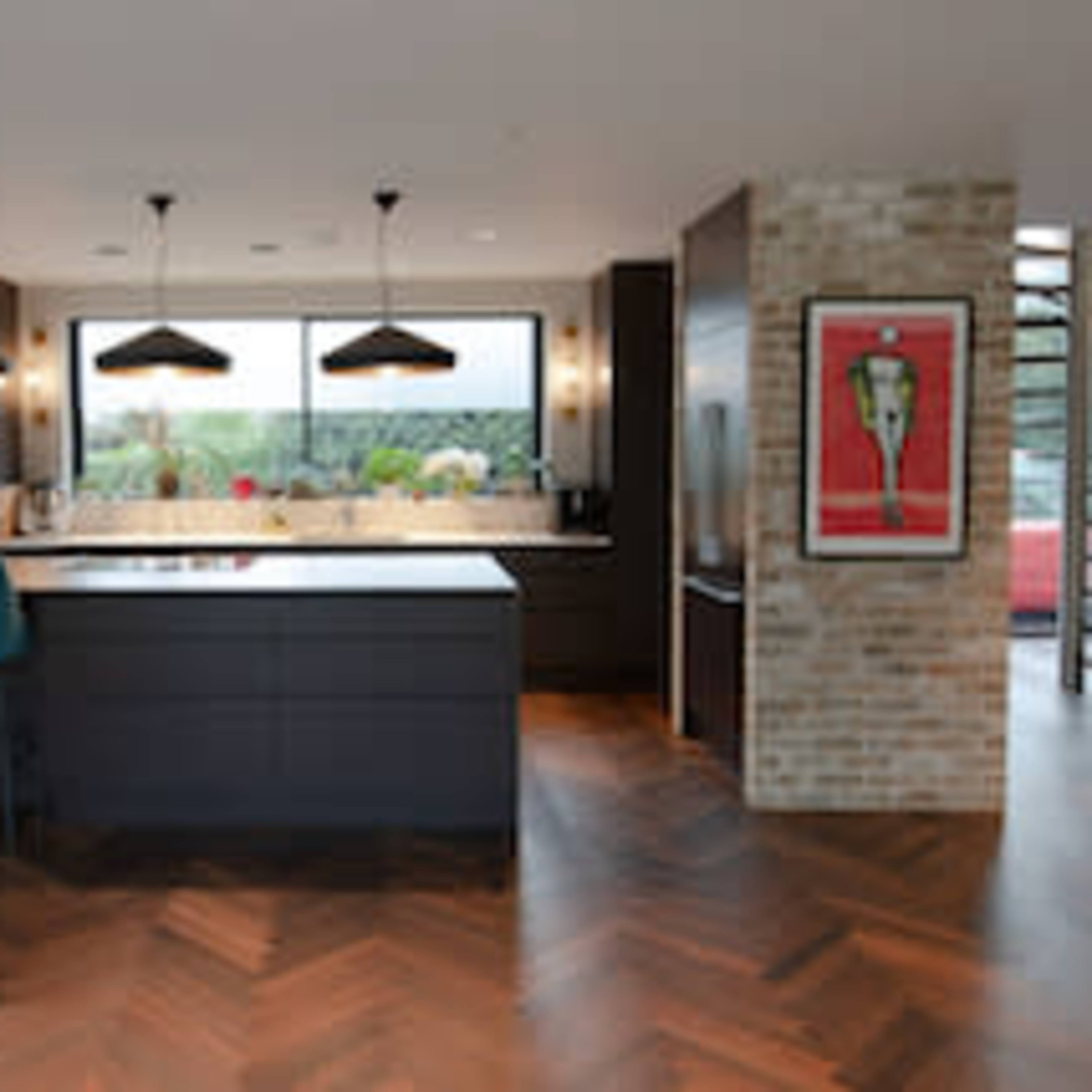 A modern kitchen features a central island with bar stools, dark cabinetry, and a large window overlooking a garden.