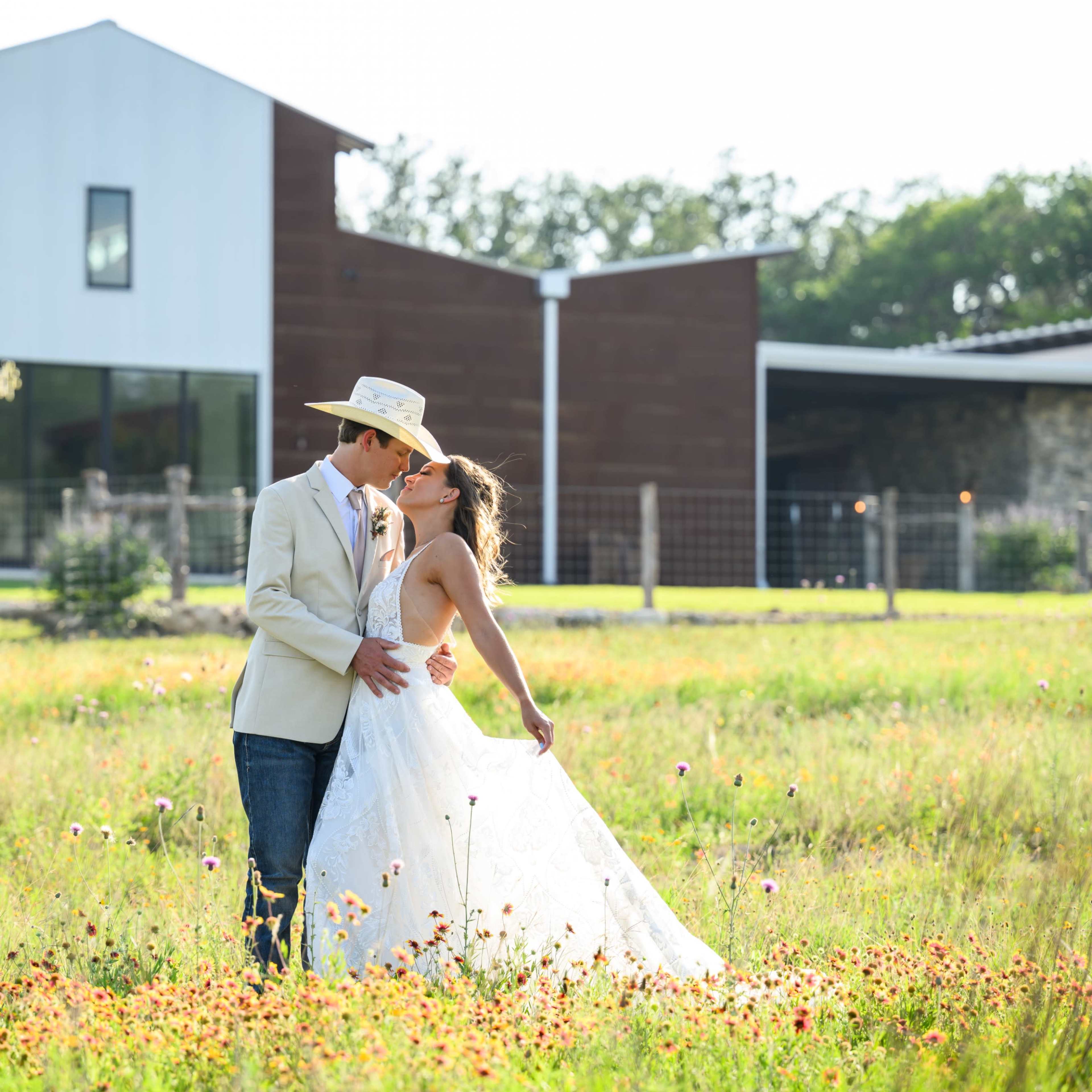 A couple in wedding attire stands in a grassy field, embracing in front of a modern building surrounded by trees.