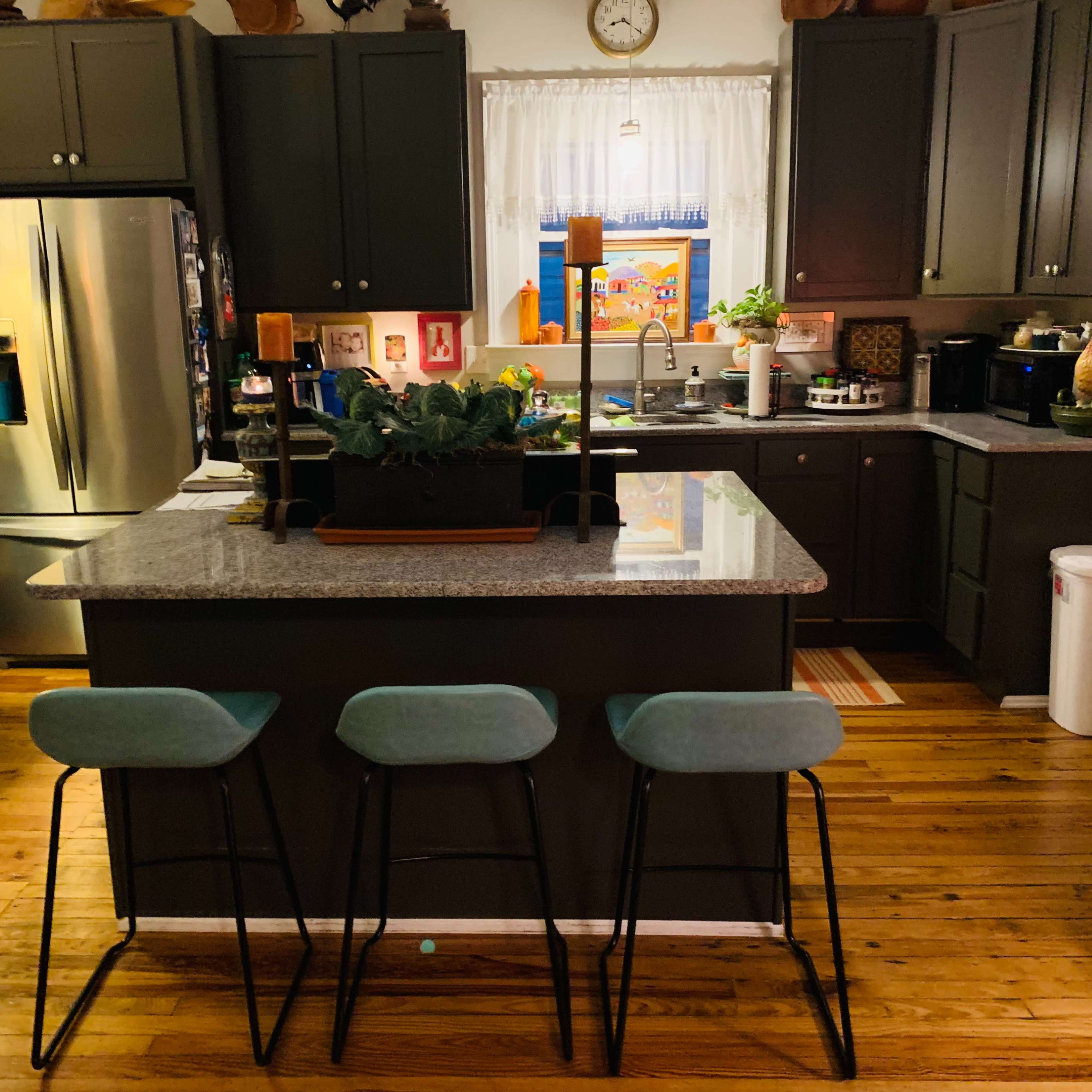 A modern kitchen with dark cabinetry, a central island with three stools, and various appliances and decorations.