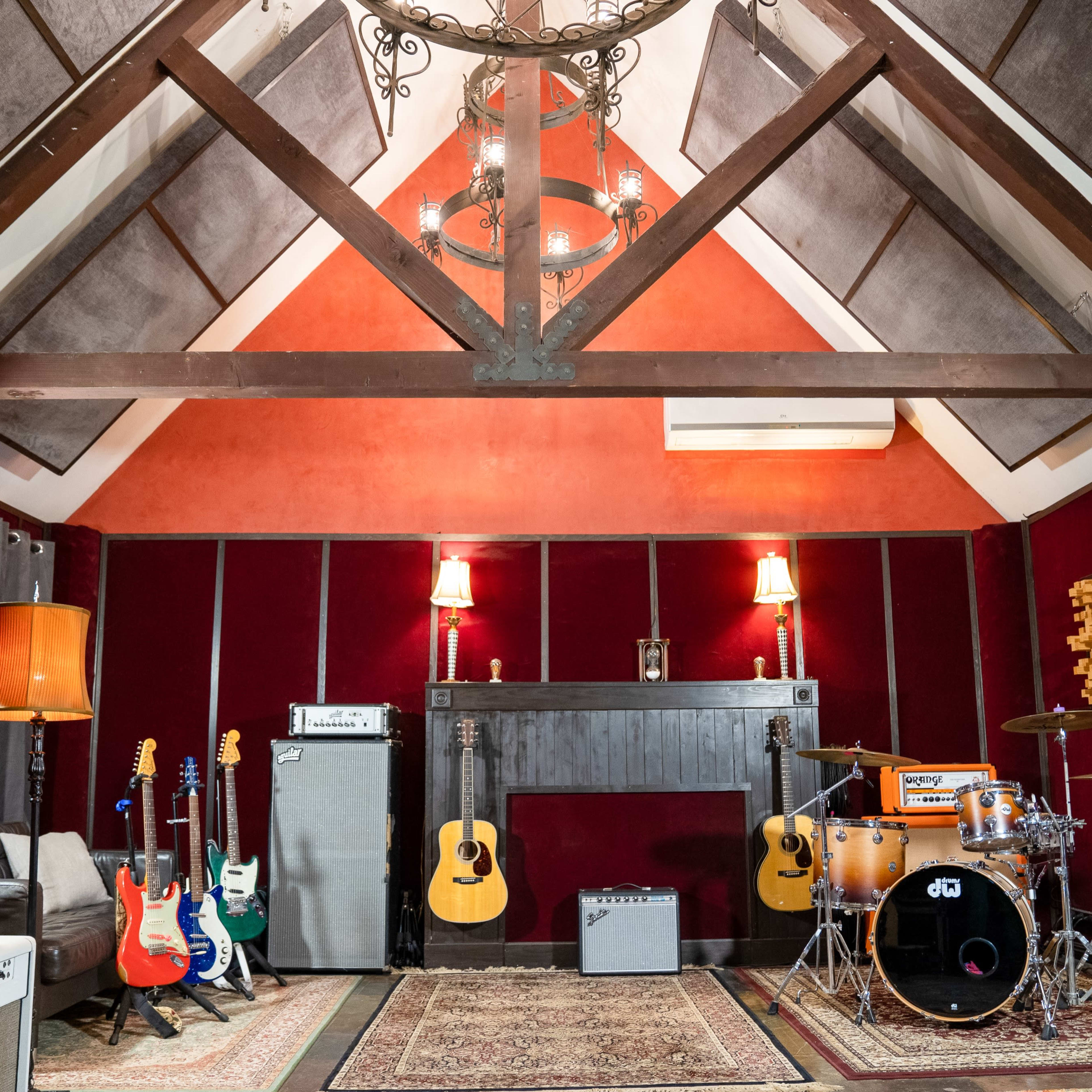 A music recording studio with various guitars, amplifiers, and a drum set arranged within a room featuring wood beams and red-paneled walls.