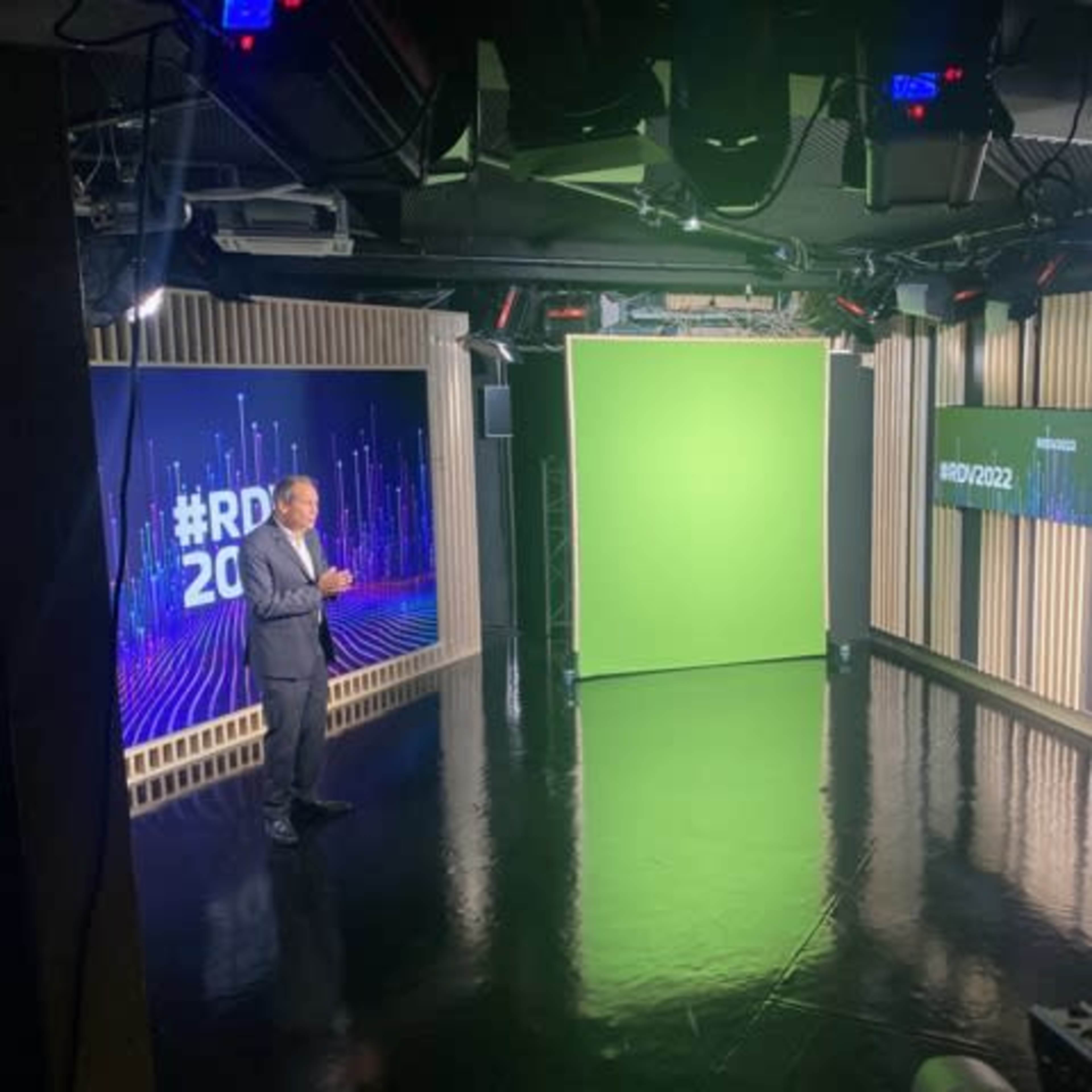 A man stands on a glossy black stage in a studio equipped with green screens and display monitors.