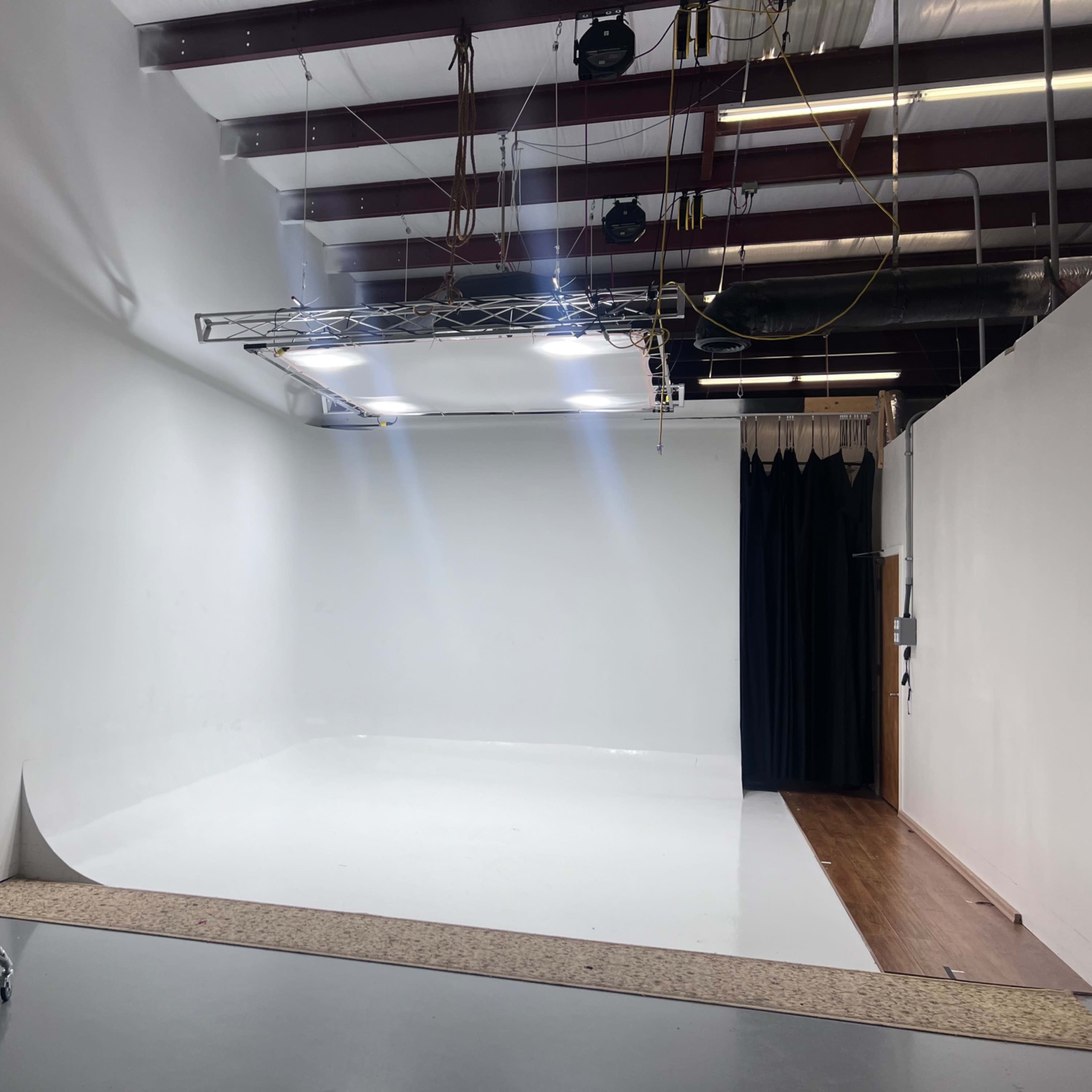 A photo studio features a large white backdrop, bright lights mounted on a truss, and black curtains along one side.