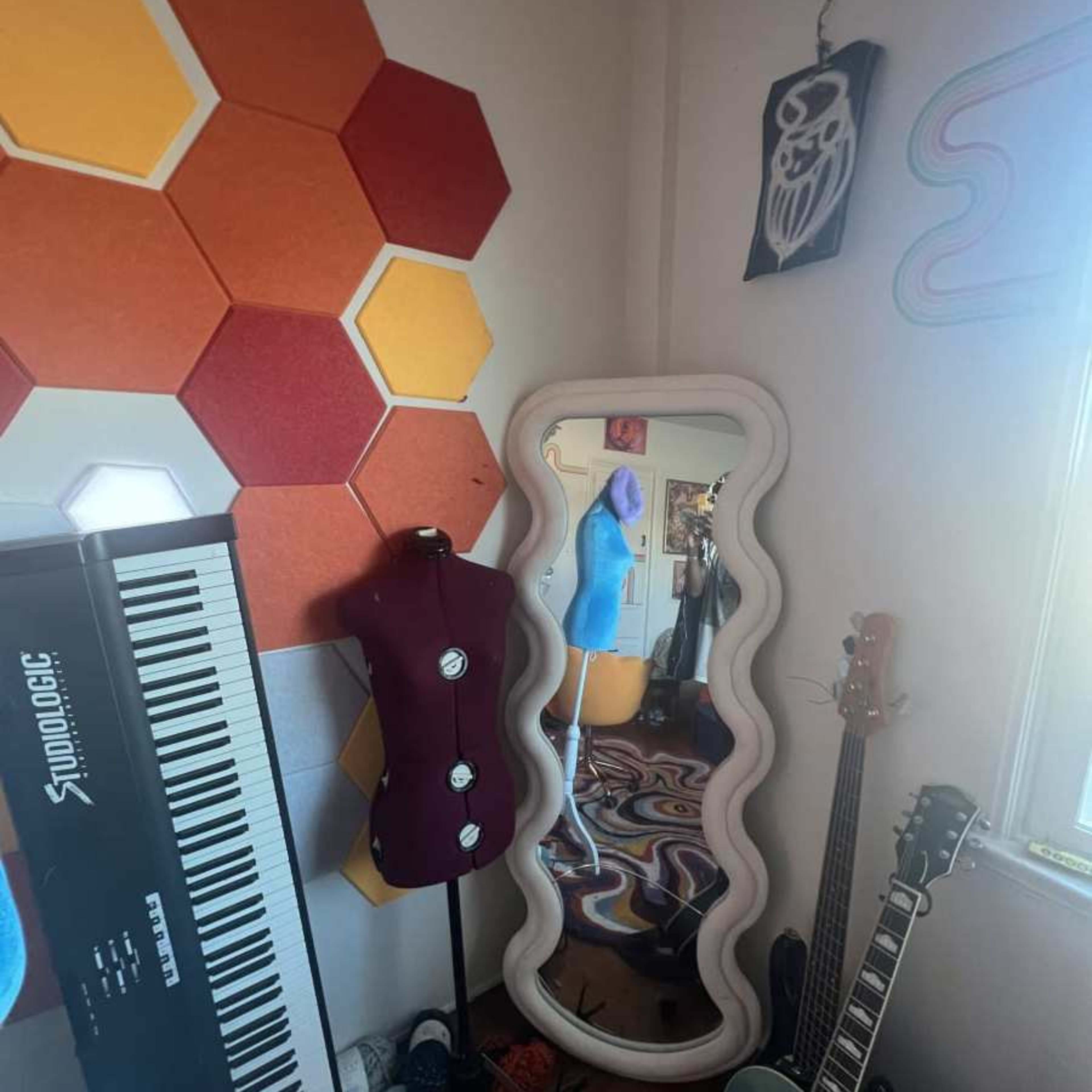 A corner of a room featuring hexagonal wall panels in orange and yellow, a full-length mirror with a wavy frame, a mannequin, a keyboard, and guitars.