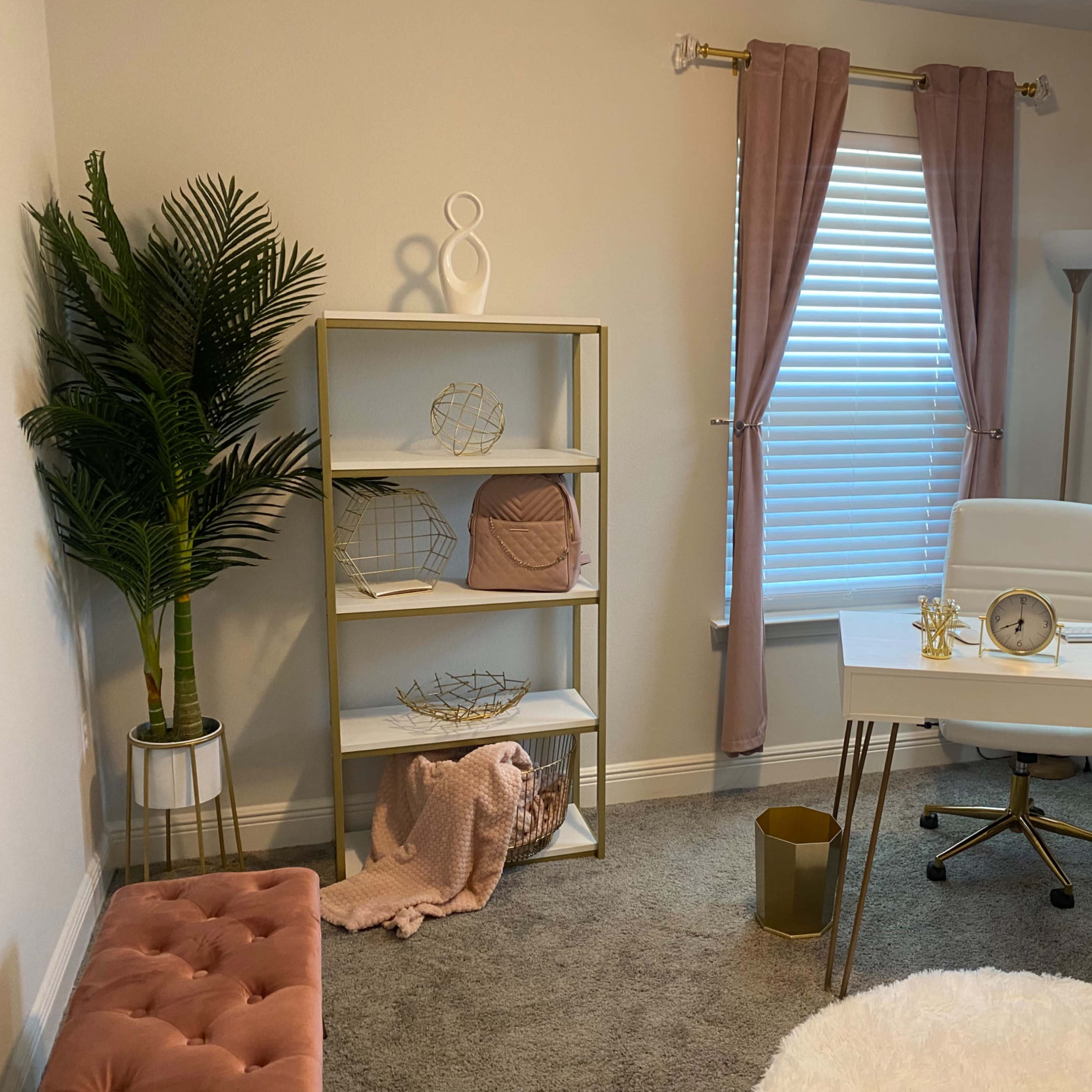 A modern home office with a white desk and chair, a tall bookshelf filled with decor, and large windows covered with pink curtains.