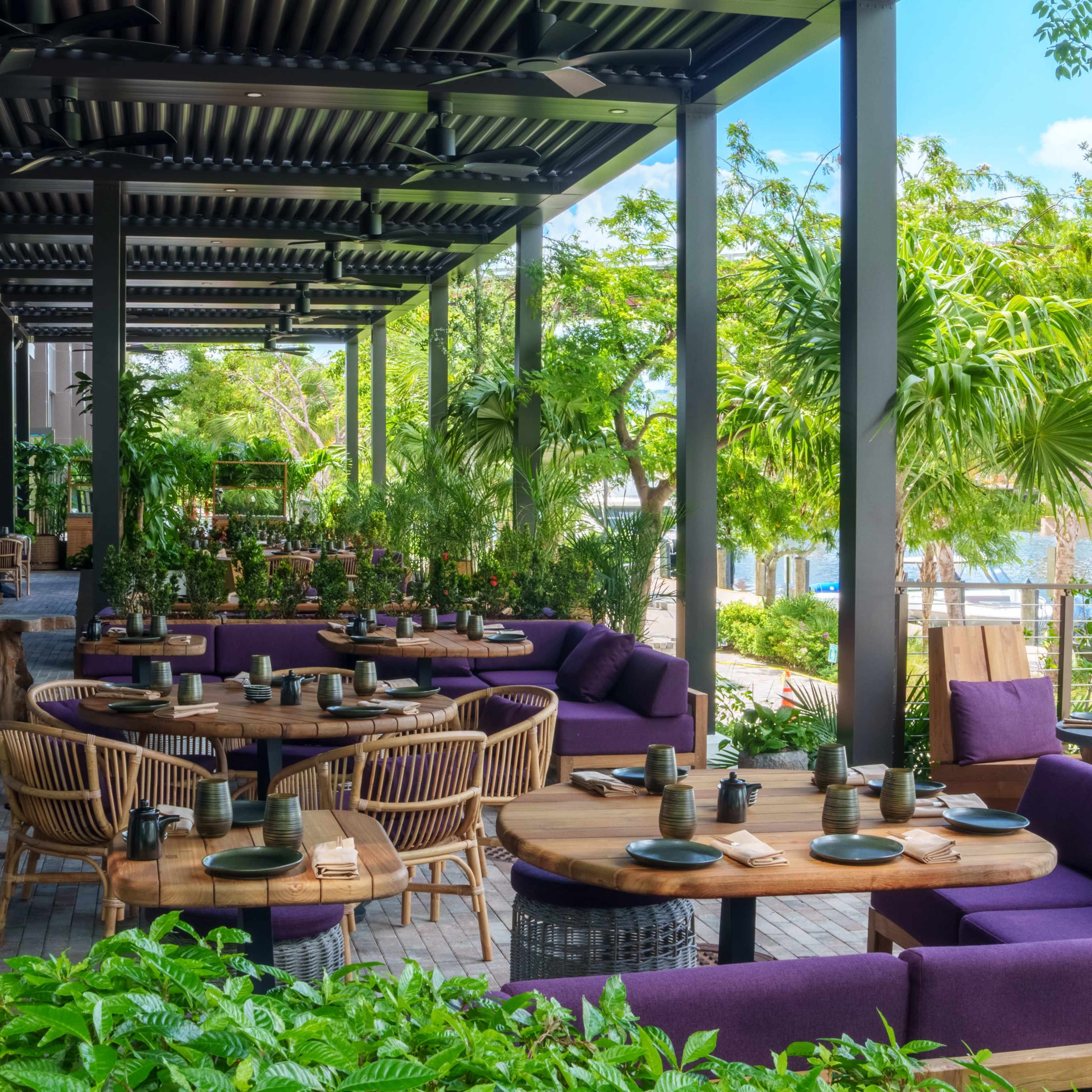 A modern restaurant patio features wooden tables surrounded by purple seating and lush greenery.