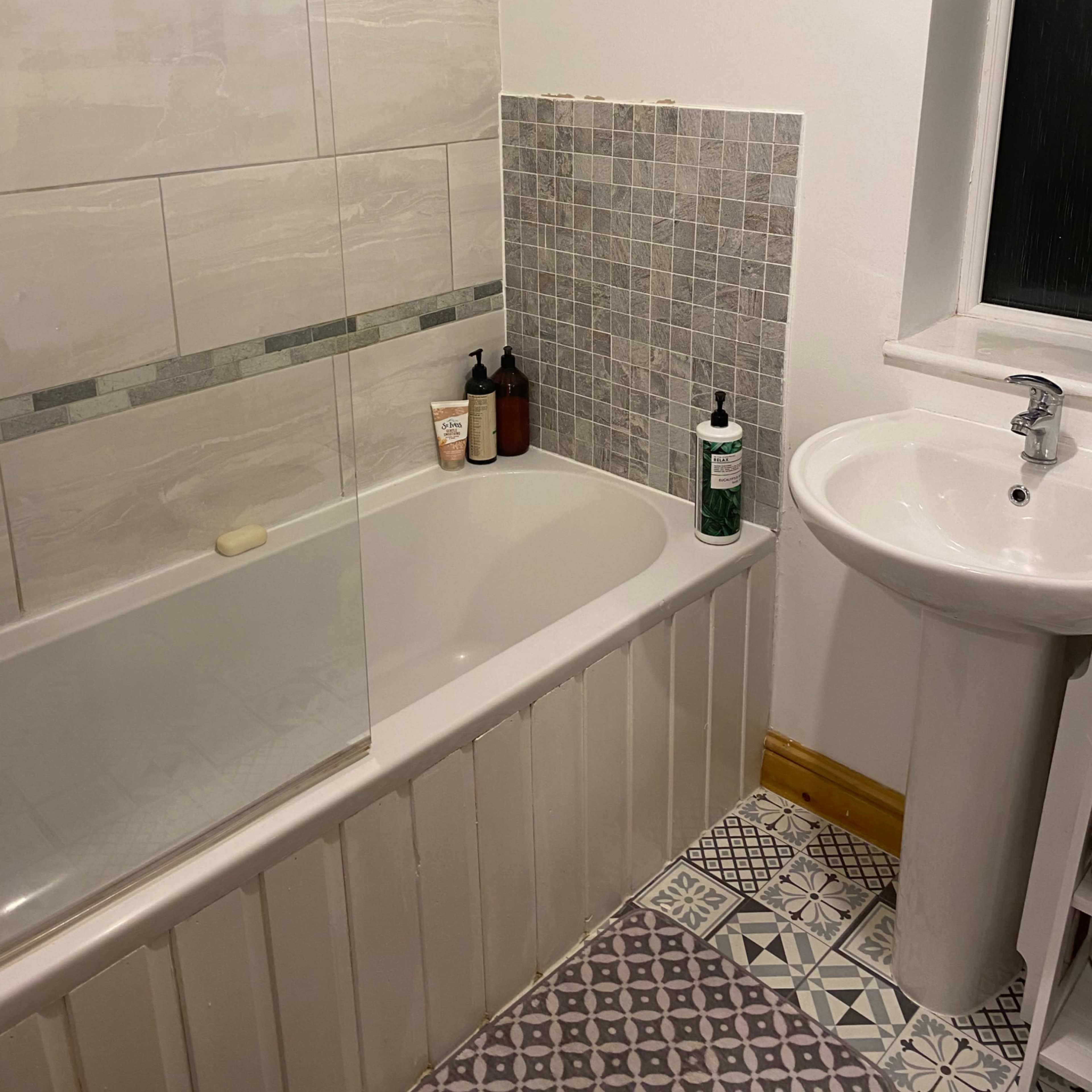 A bathroom featuring a bathtub with a glass screen, a pedestal sink, and patterned tiles on the floor.