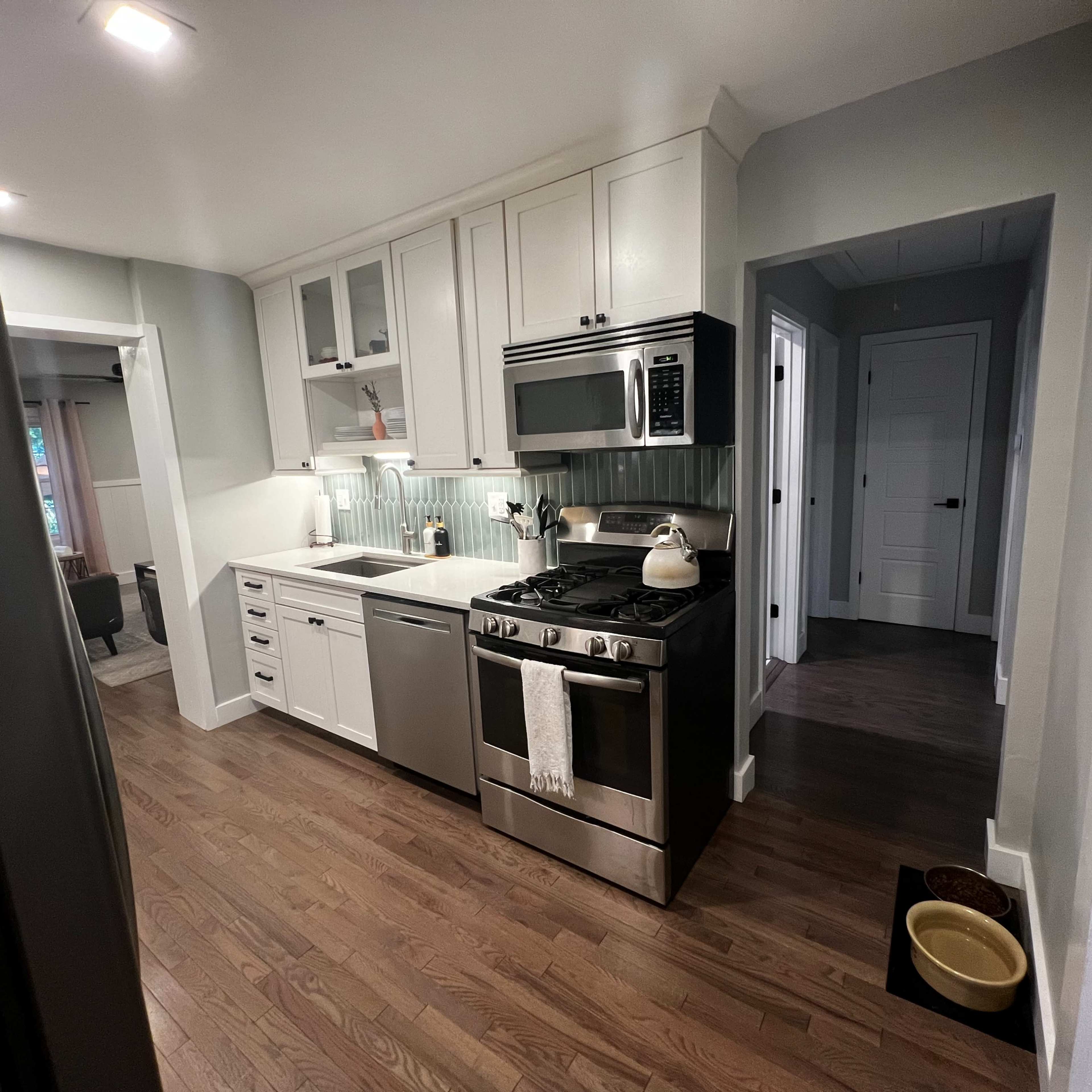 A modern kitchen with white cabinets, stainless steel appliances, and hardwood flooring, adjacent to a hallway.
