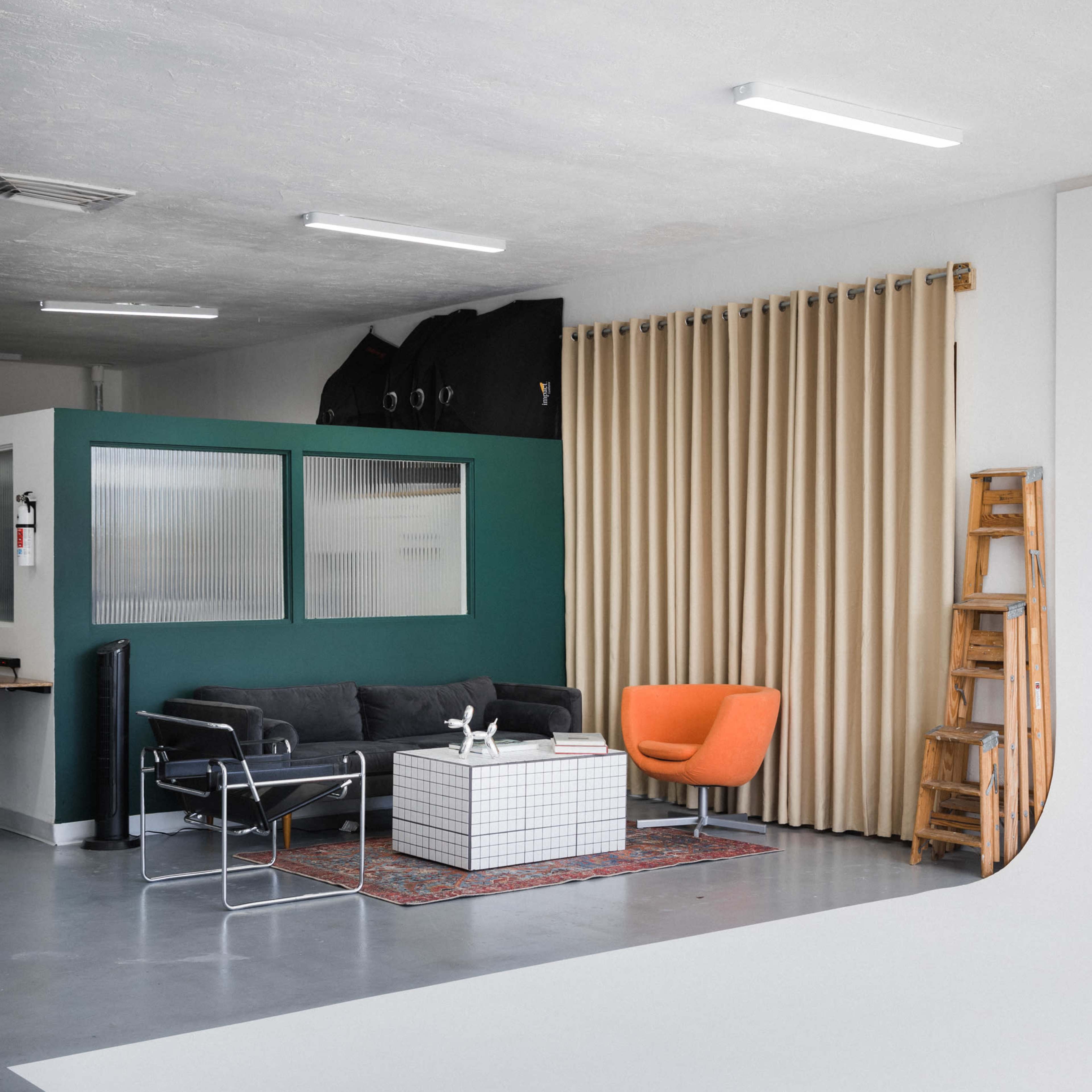 A modern interior space featuring a black sofa, an orange armchair, a white coffee table, and a wooden ladder against a backdrop of green walls and beige curtains.
