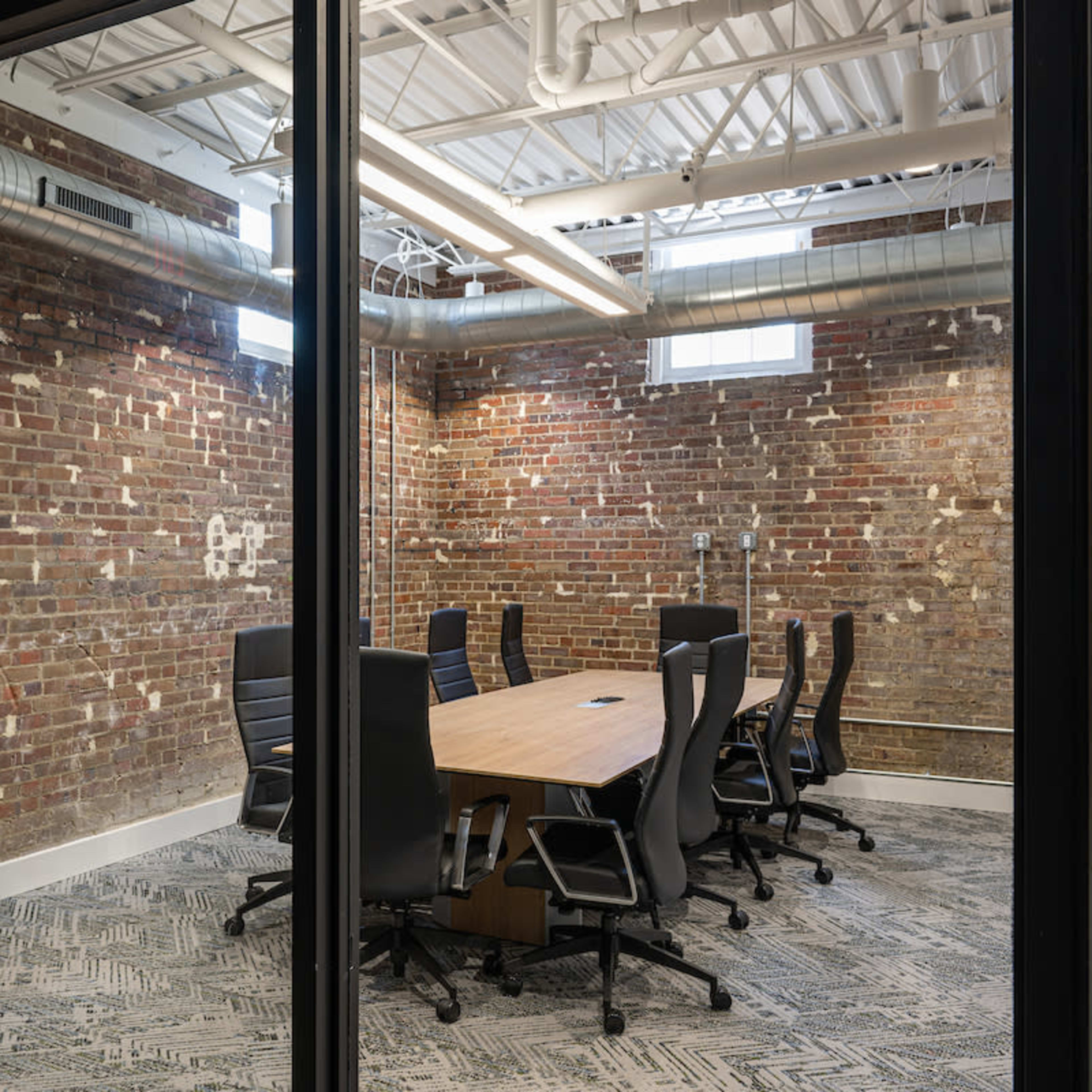 A modern conference room with a wooden table surrounded by black office chairs, set against a brick wall.