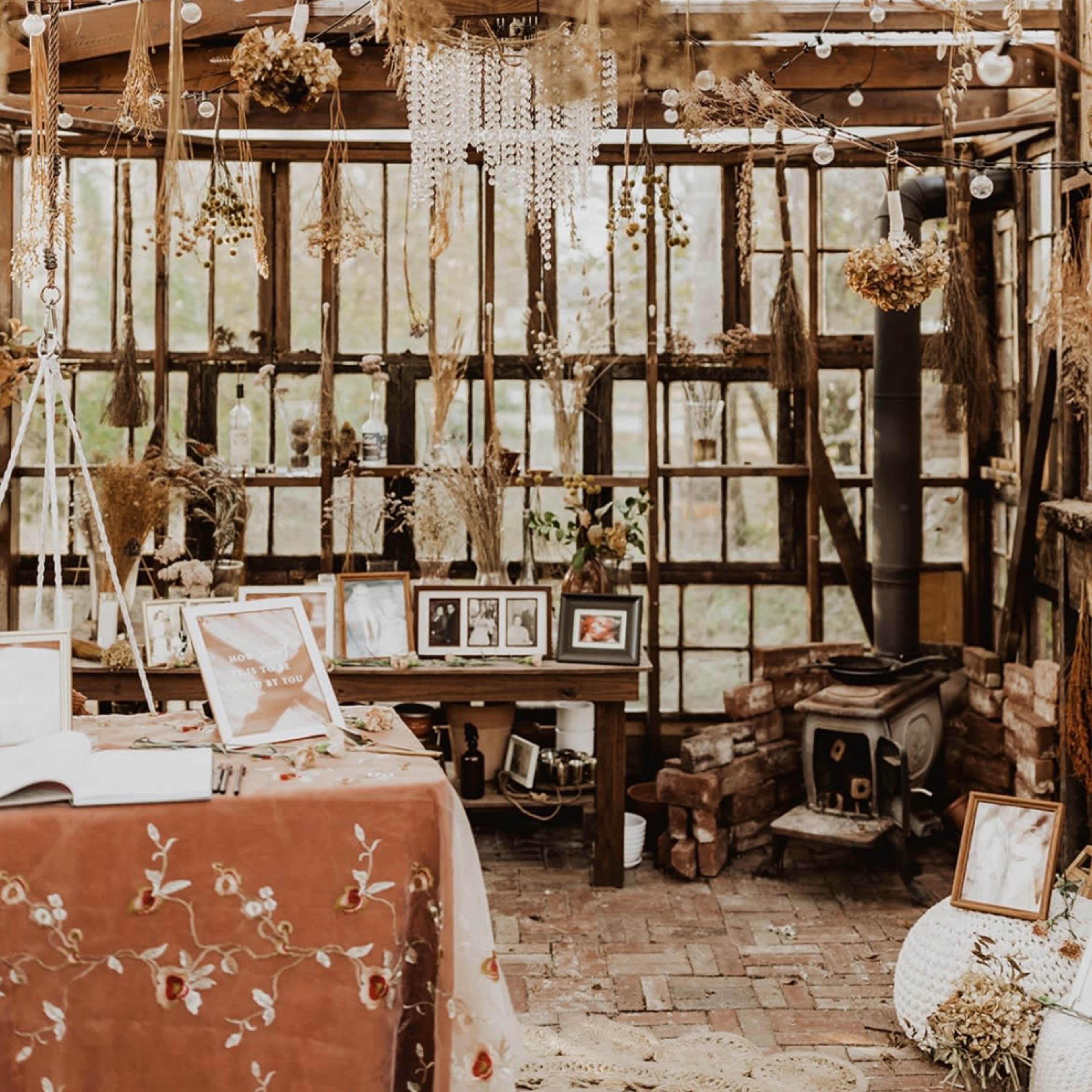 A rustic indoor space filled with hanging dried flowers and decorative elements, featuring a table with photographs in front of a windowed wall.