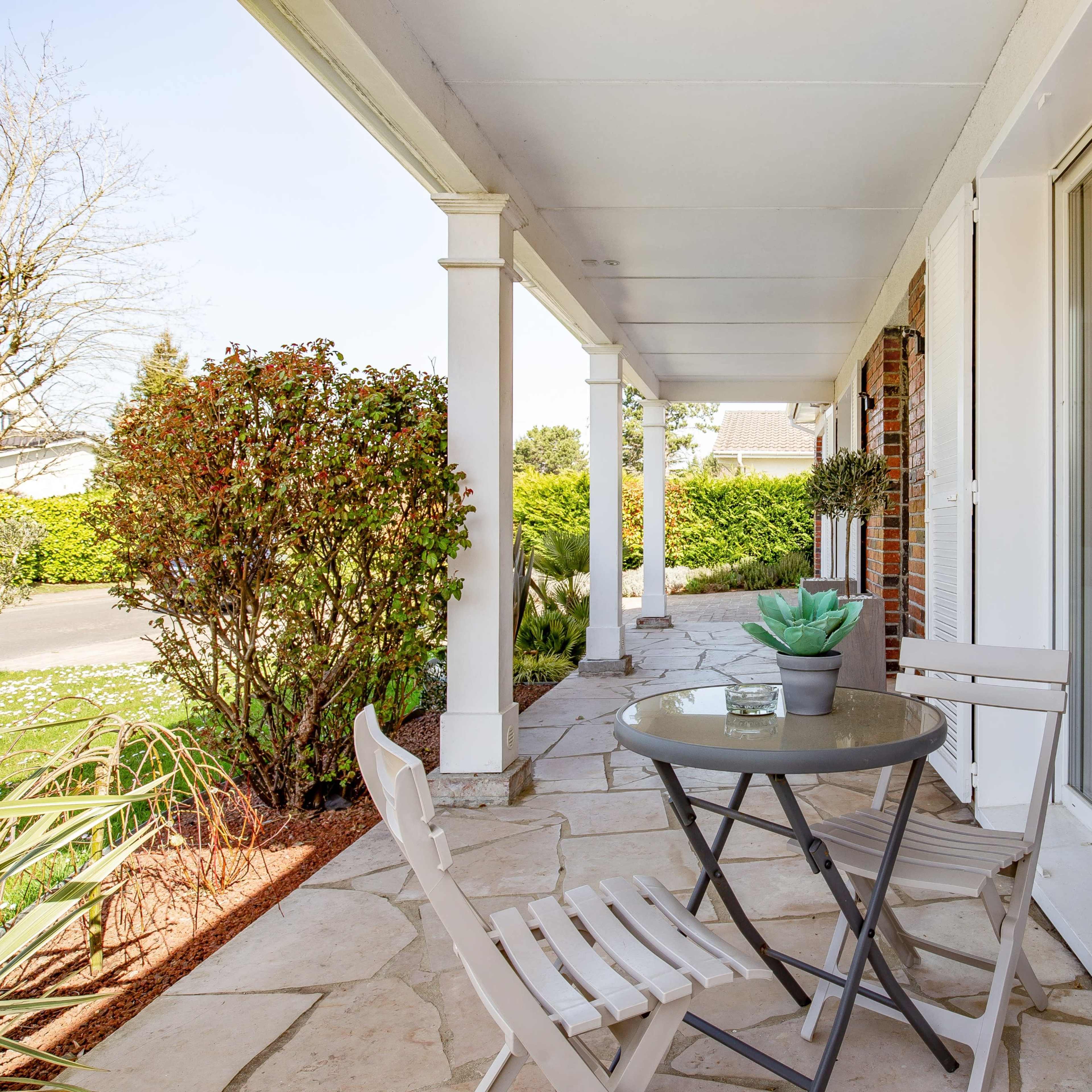 A small patio features a round table with two chairs, surrounded by shrubbery and a view of a quiet street.