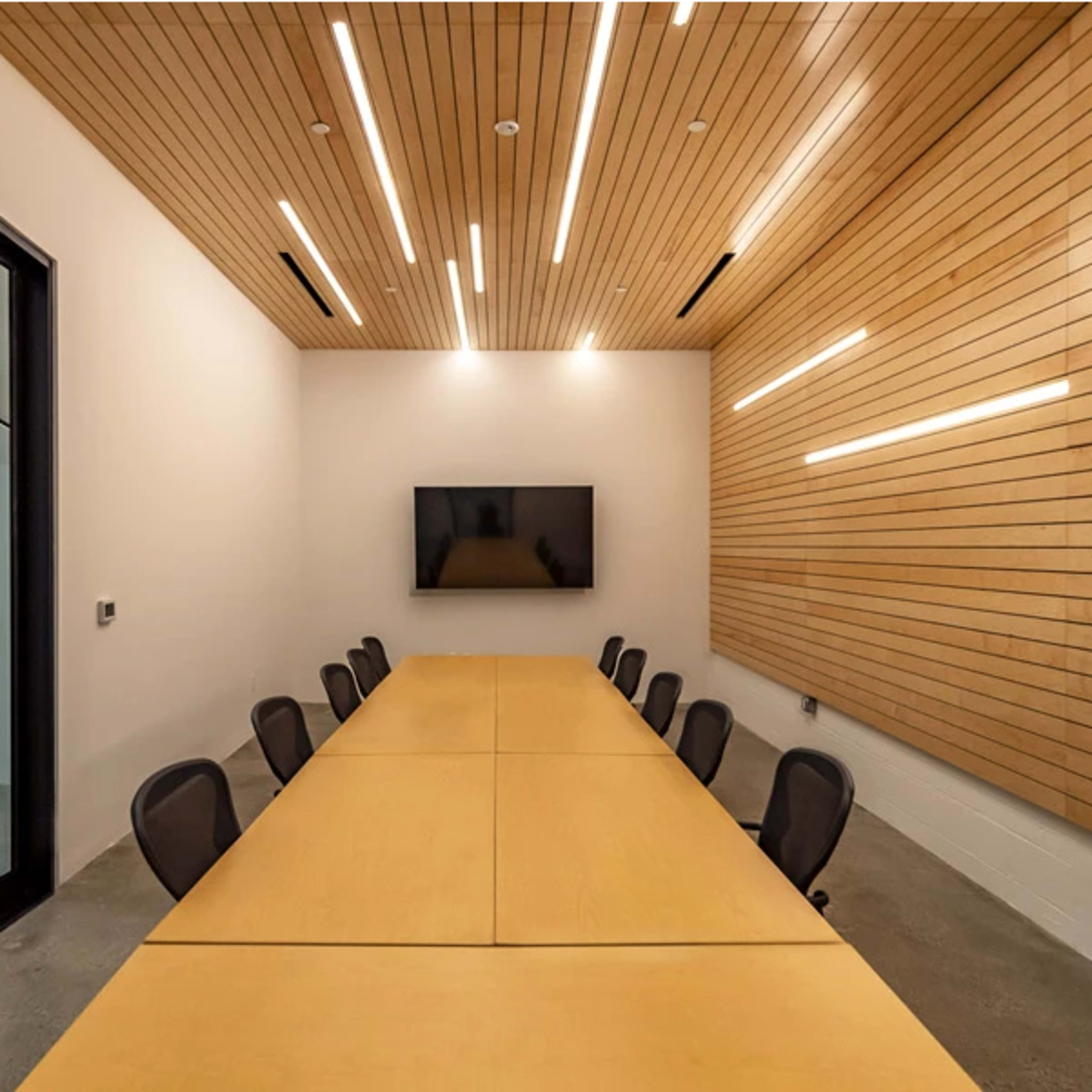 A modern conference room with a long wooden table, black chairs, and a large screen on the wall, illuminated by linear ceiling lights.