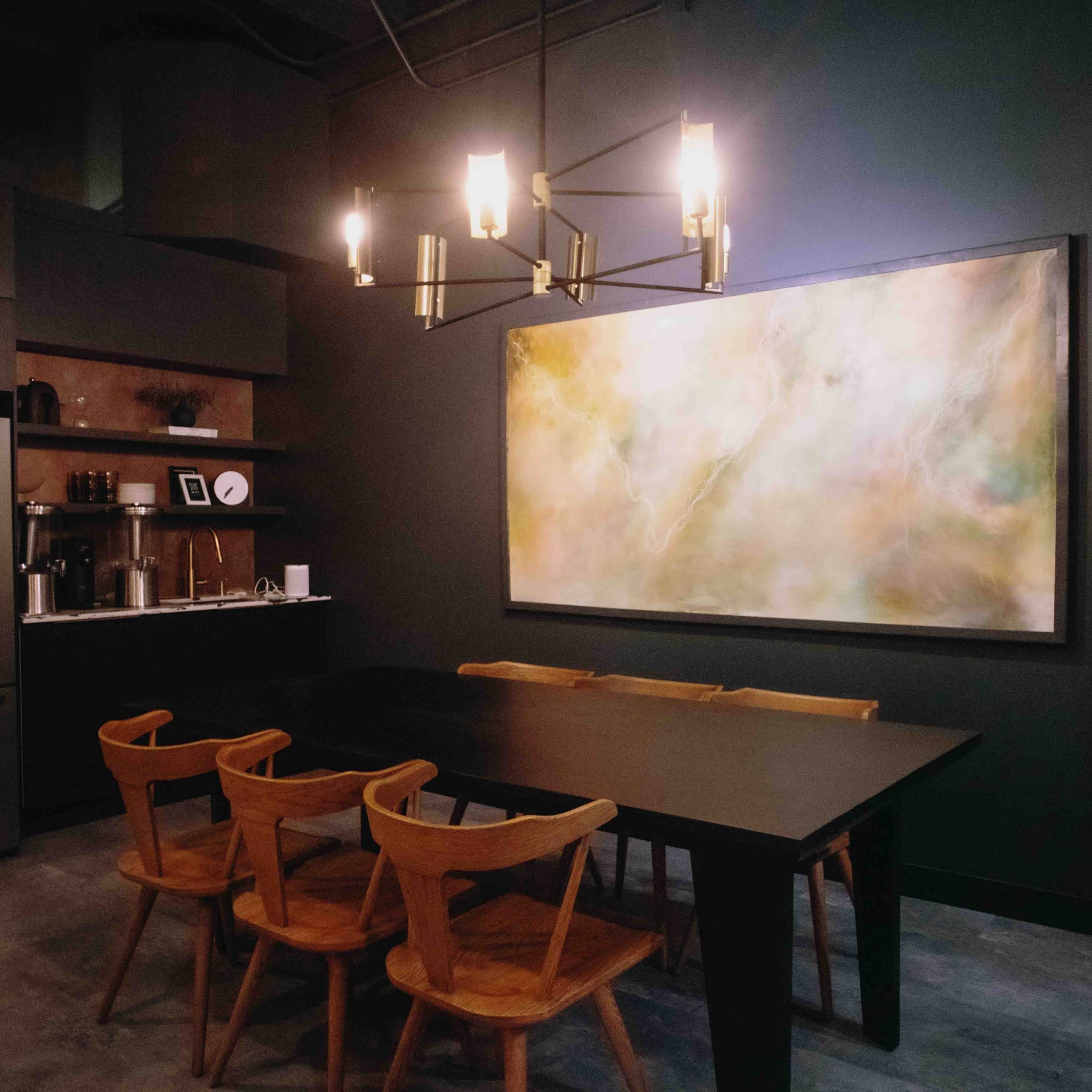 A dining area features a large black table surrounded by wooden chairs, an abstract painting on the wall, and a modern kitchen setup with stainless steel appliances.