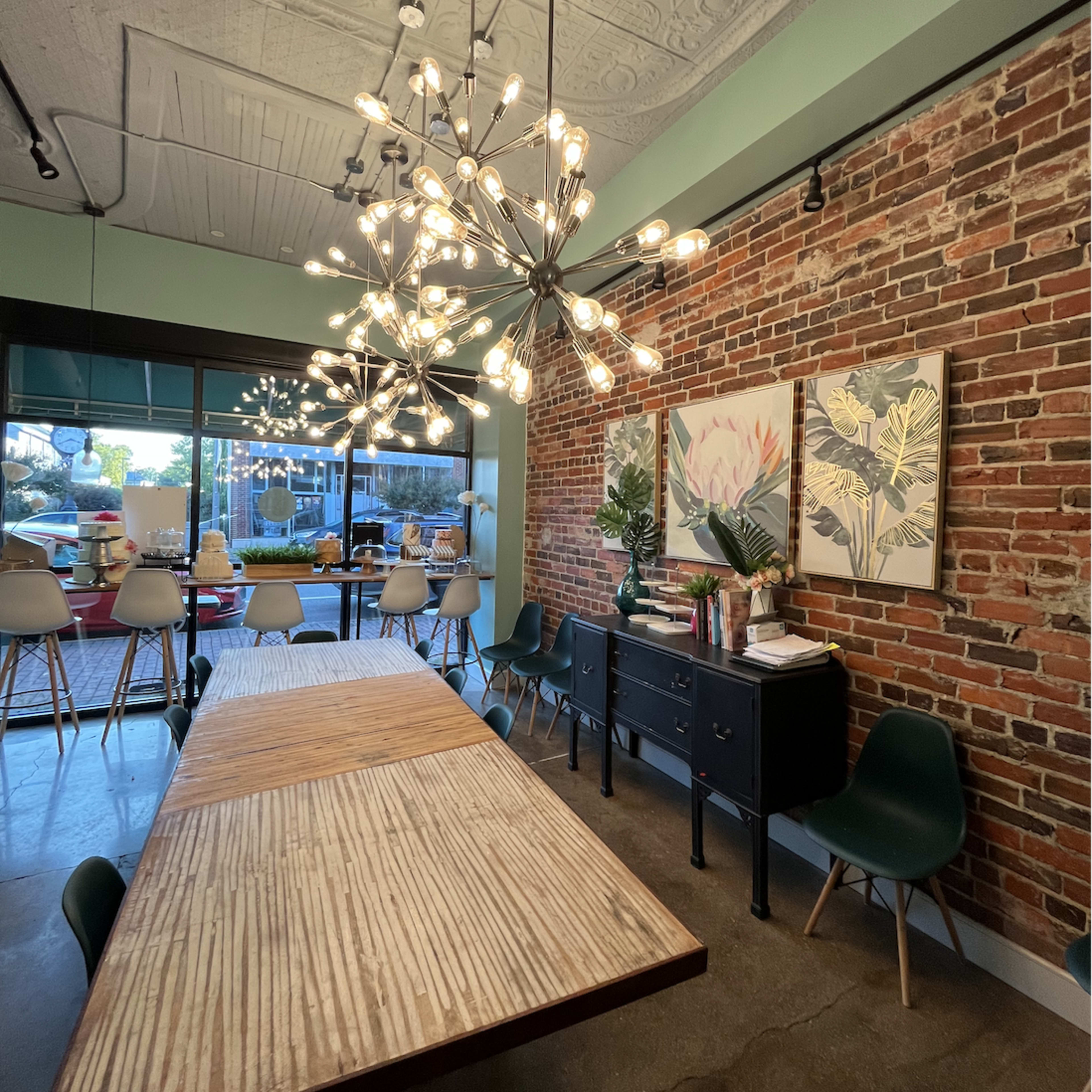 A dining area with a large wooden table, modern seating, a decorative cabinet, and two pieces of framed artwork beneath two contemporary light fixtures, all set against a brick wall.