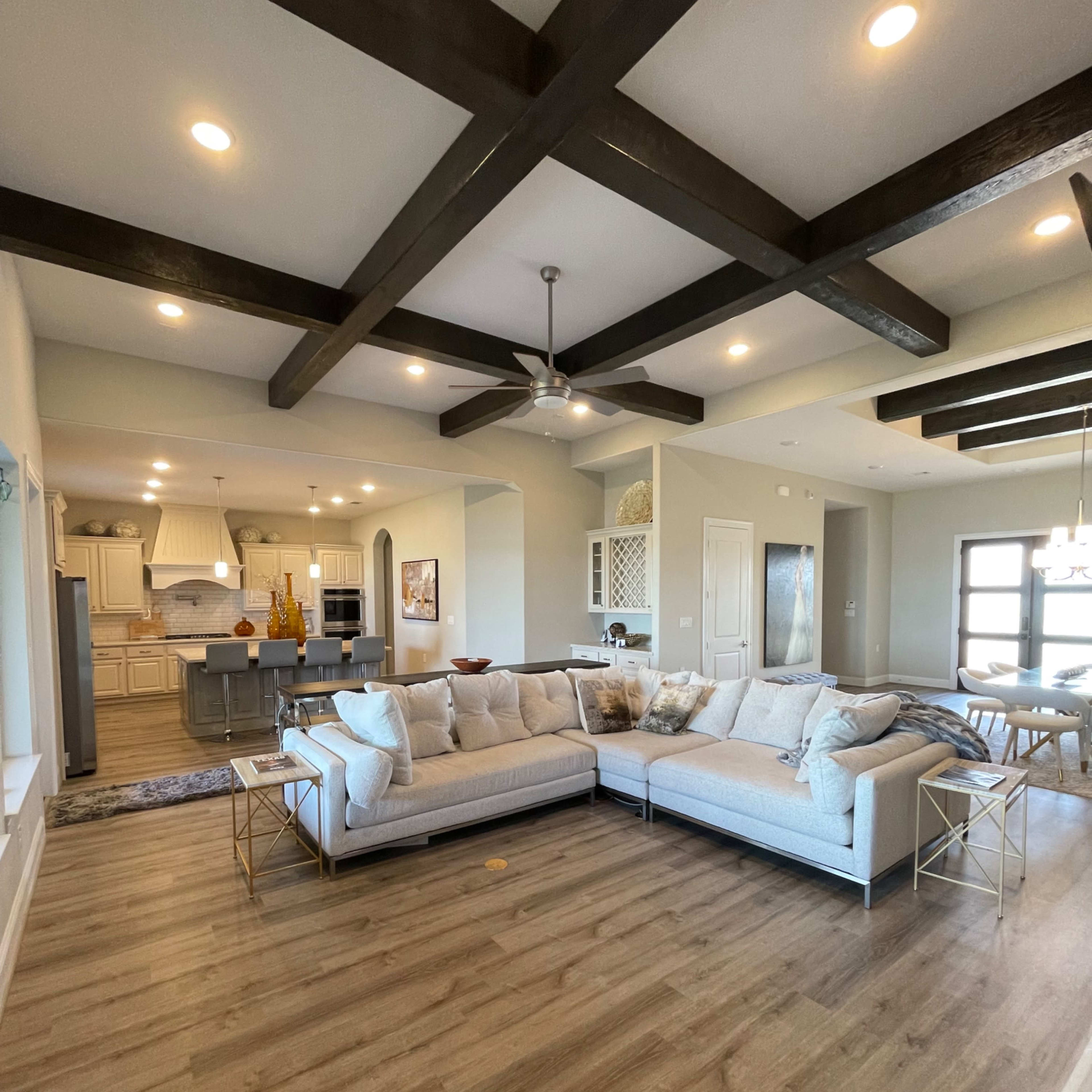 A spacious living room features a large sectional sofa, wooden beams on the ceiling, and an open layout connecting to a kitchen area.
