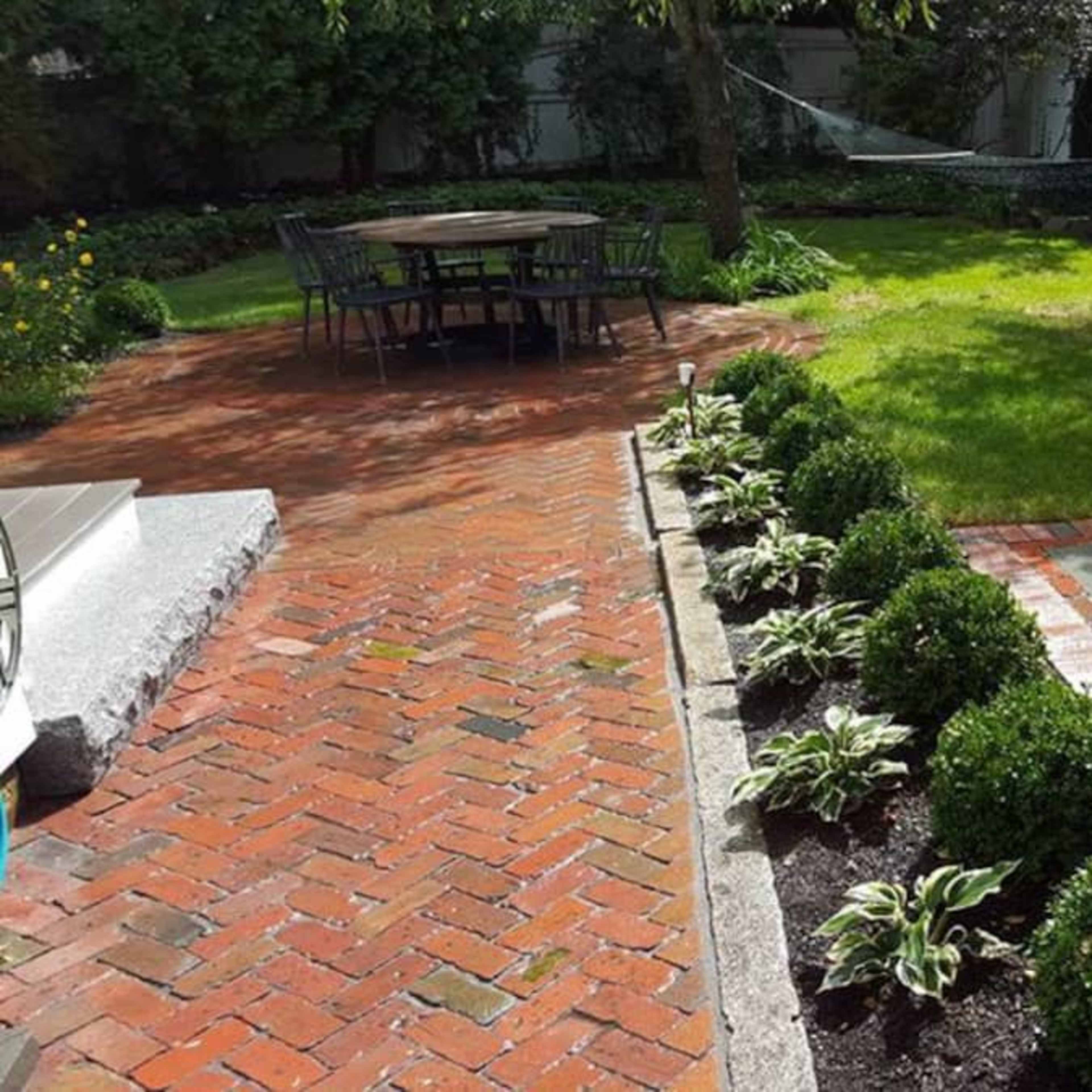 A backyard featuring a brick pathway, a dining area with a table and chairs, a hammock, a grassy lawn, and a modern fire pit surrounded by neatly trimmed bushes.