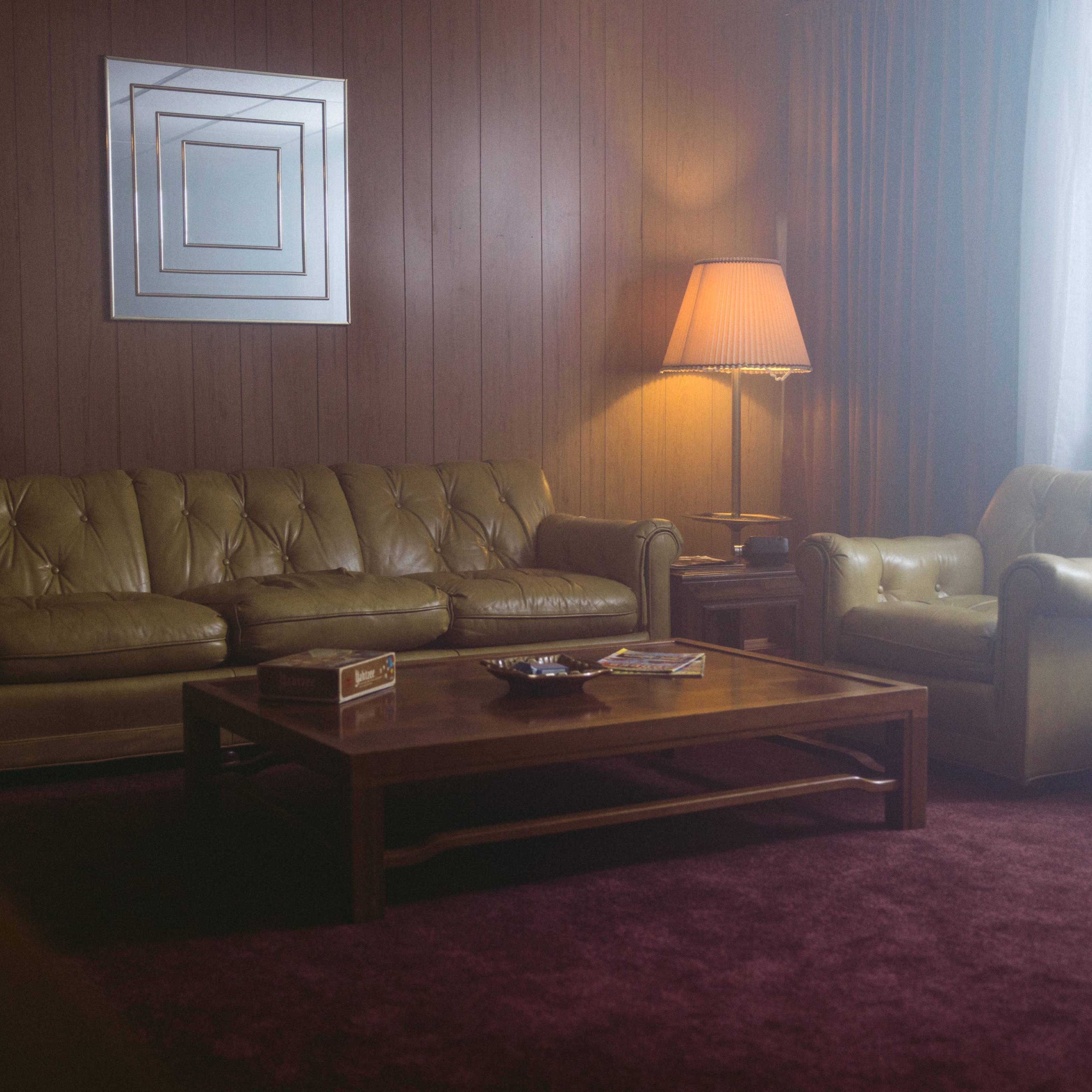 A dimly lit living room featuring a yellow leather couch, a matching armchair, a wooden coffee table, and a floor lamp beside a large window with sheer curtains.