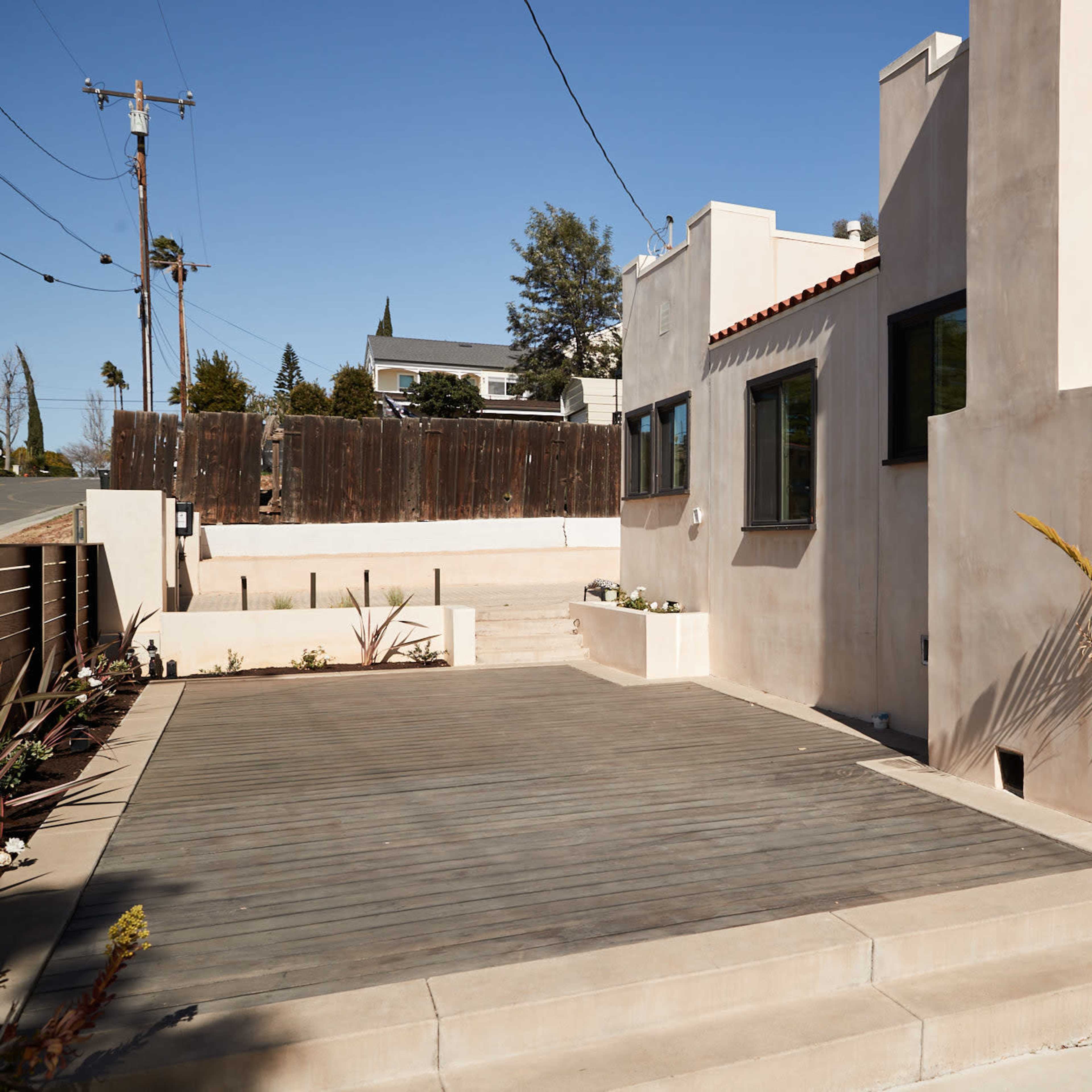 A modern, minimalist outdoor patio area with wooden decking and a potted plant, adjacent to a light-colored building.