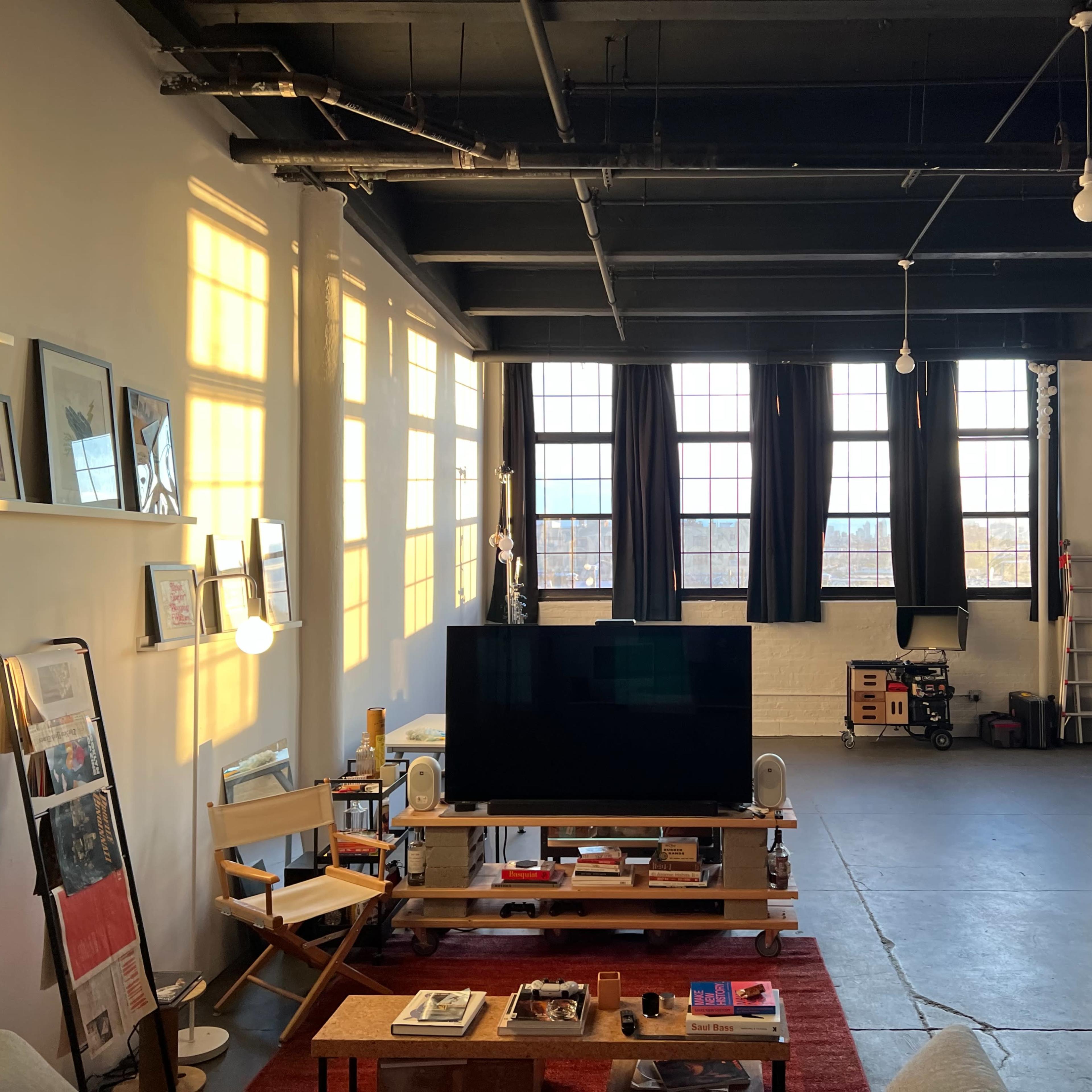 A spacious loft-like living area with large windows, a television on a wooden table, and various furniture arranged in an open layout.