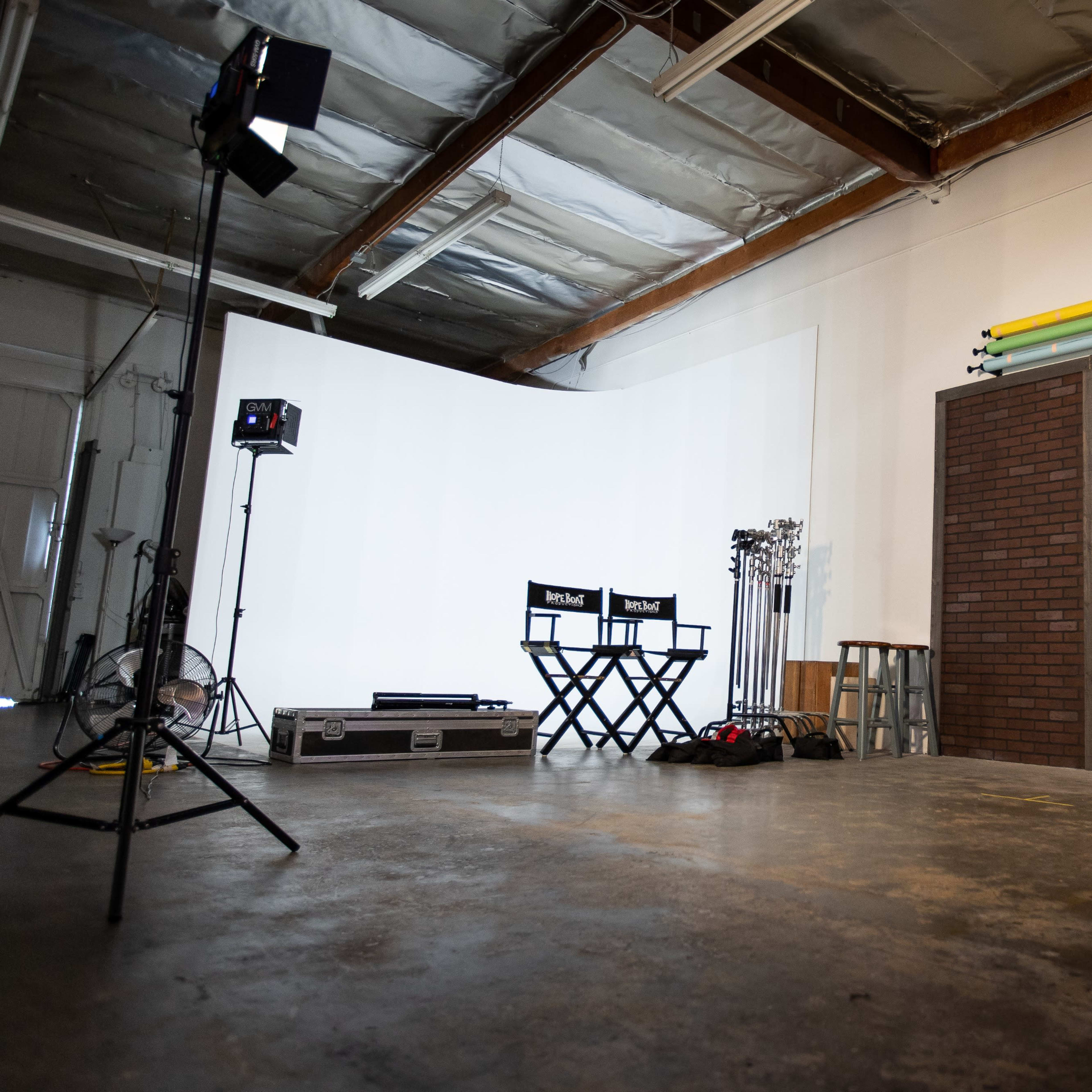 A photography studio featuring two director's chairs, a backdrop with a brick design, and lighting equipment set up against a white wall.