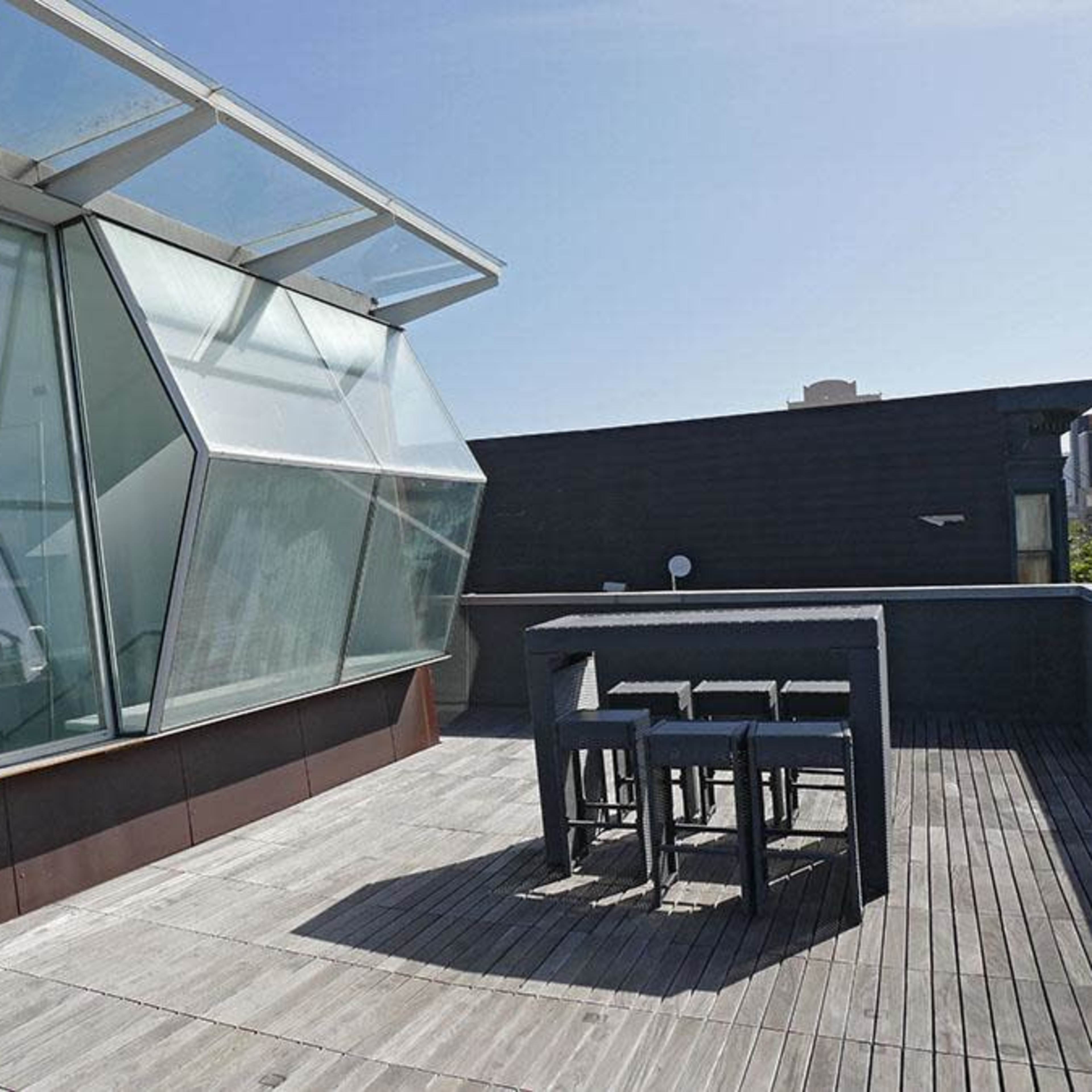 A modern rooftop terrace features a geometric glass structure and a wooden deck with a dining table and chairs.