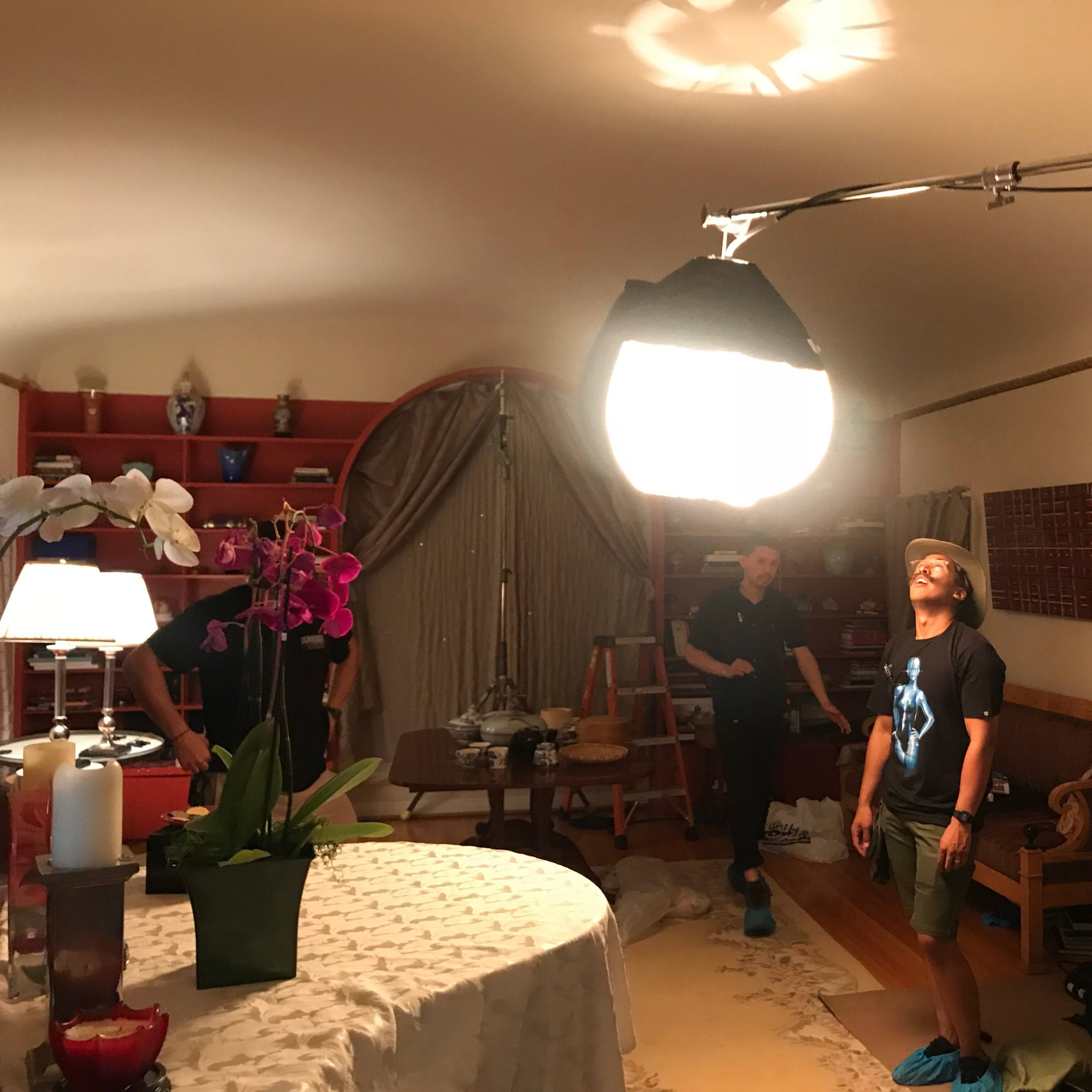 A film crew sets up lighting equipment in a living room, with one crew member looking up at a large light fixture.