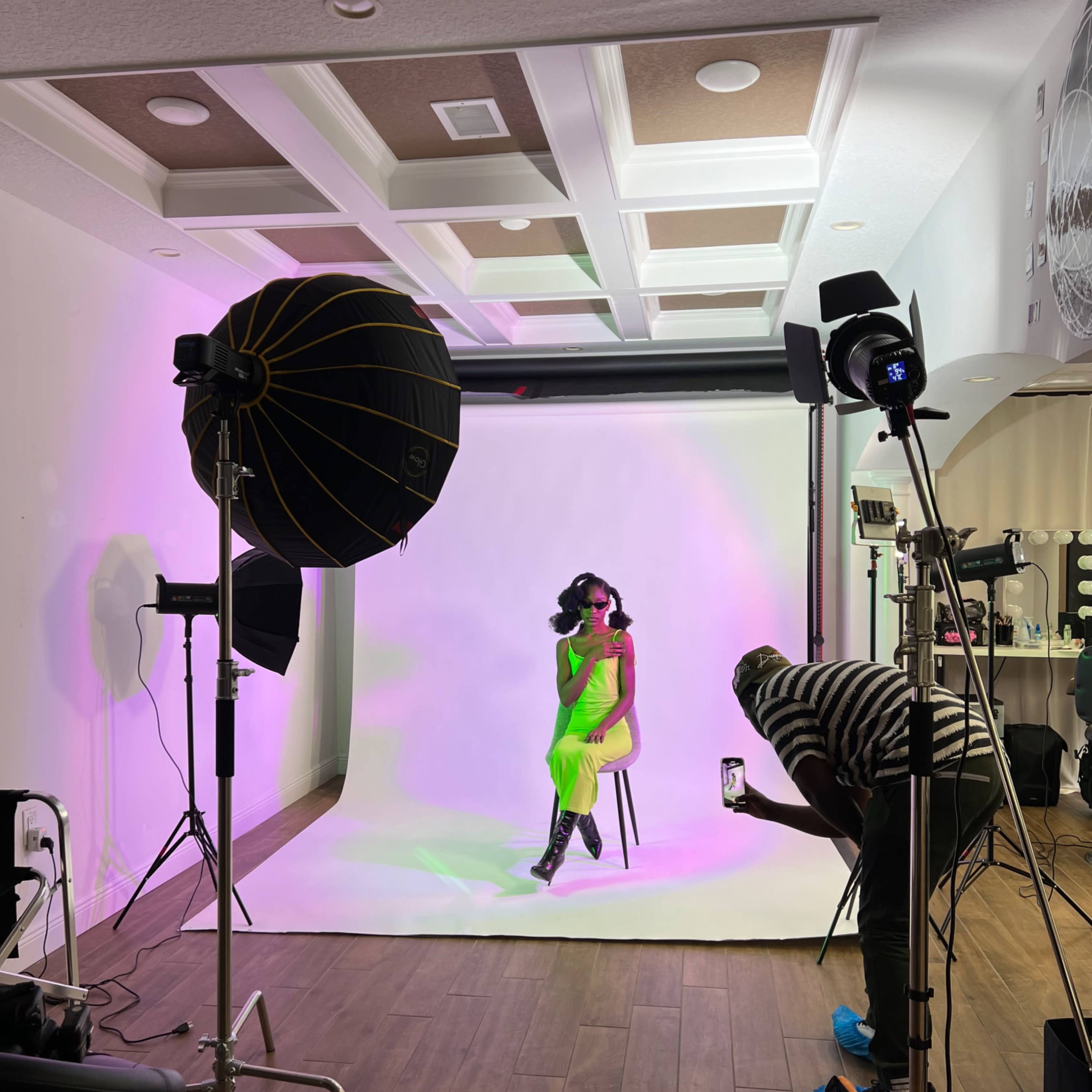 A model in a bright green outfit sits on a chair in a photography studio, surrounded by lighting equipment and a backdrop.