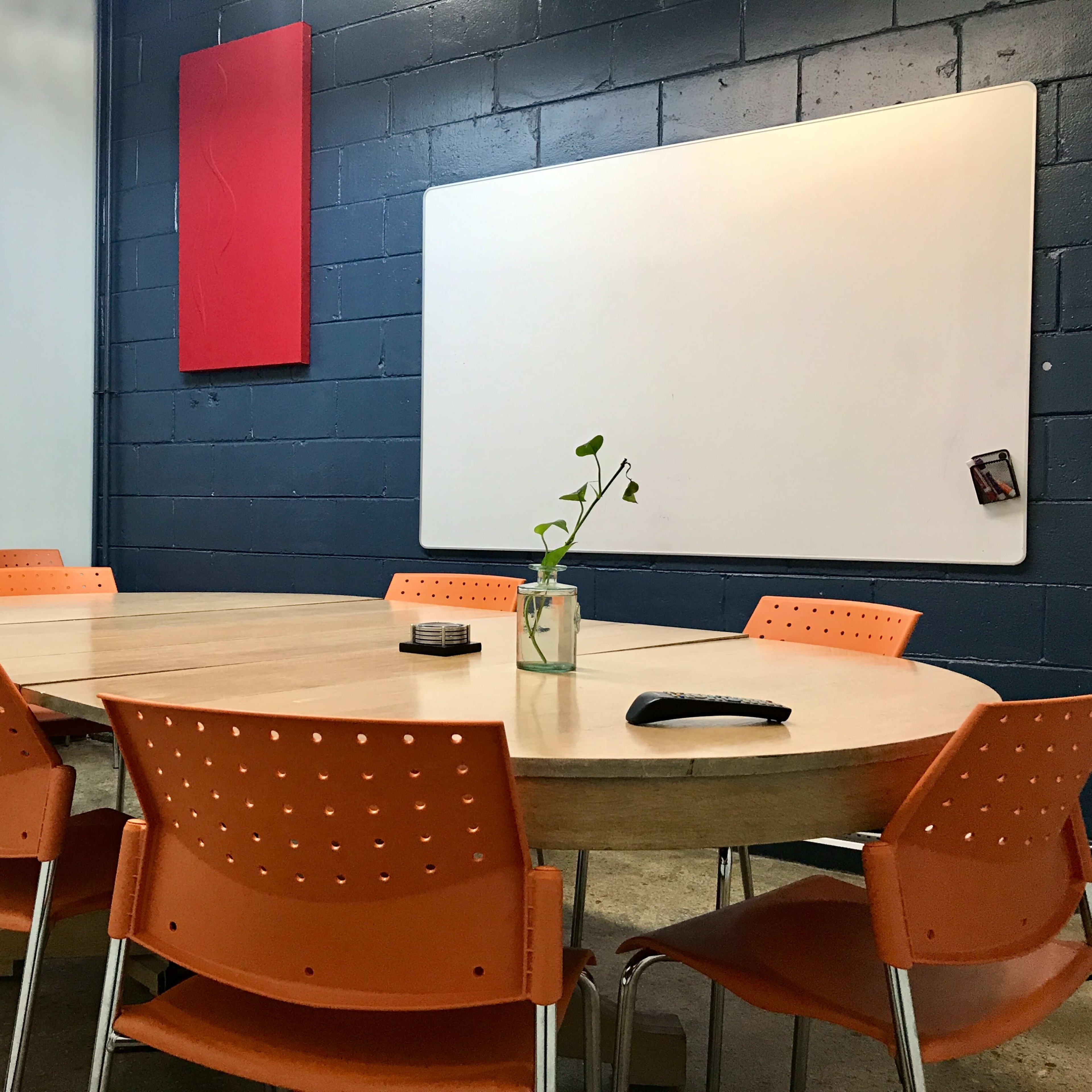 A round wooden table surrounded by eight orange chairs is set in a room with a blue wall and a whiteboard.