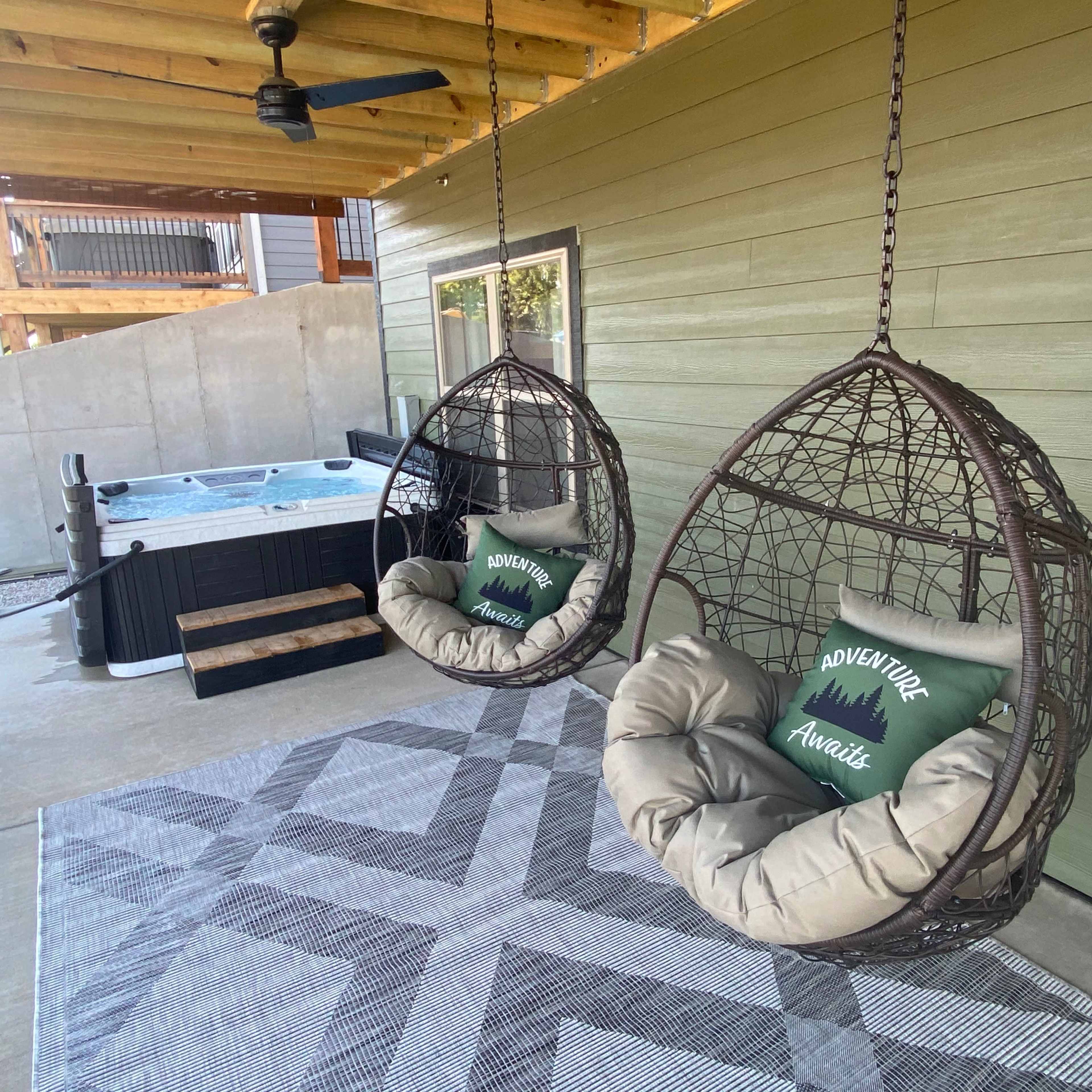 Two hanging wicker chairs with cushions next to a hot tub on a patio with wooden beams.