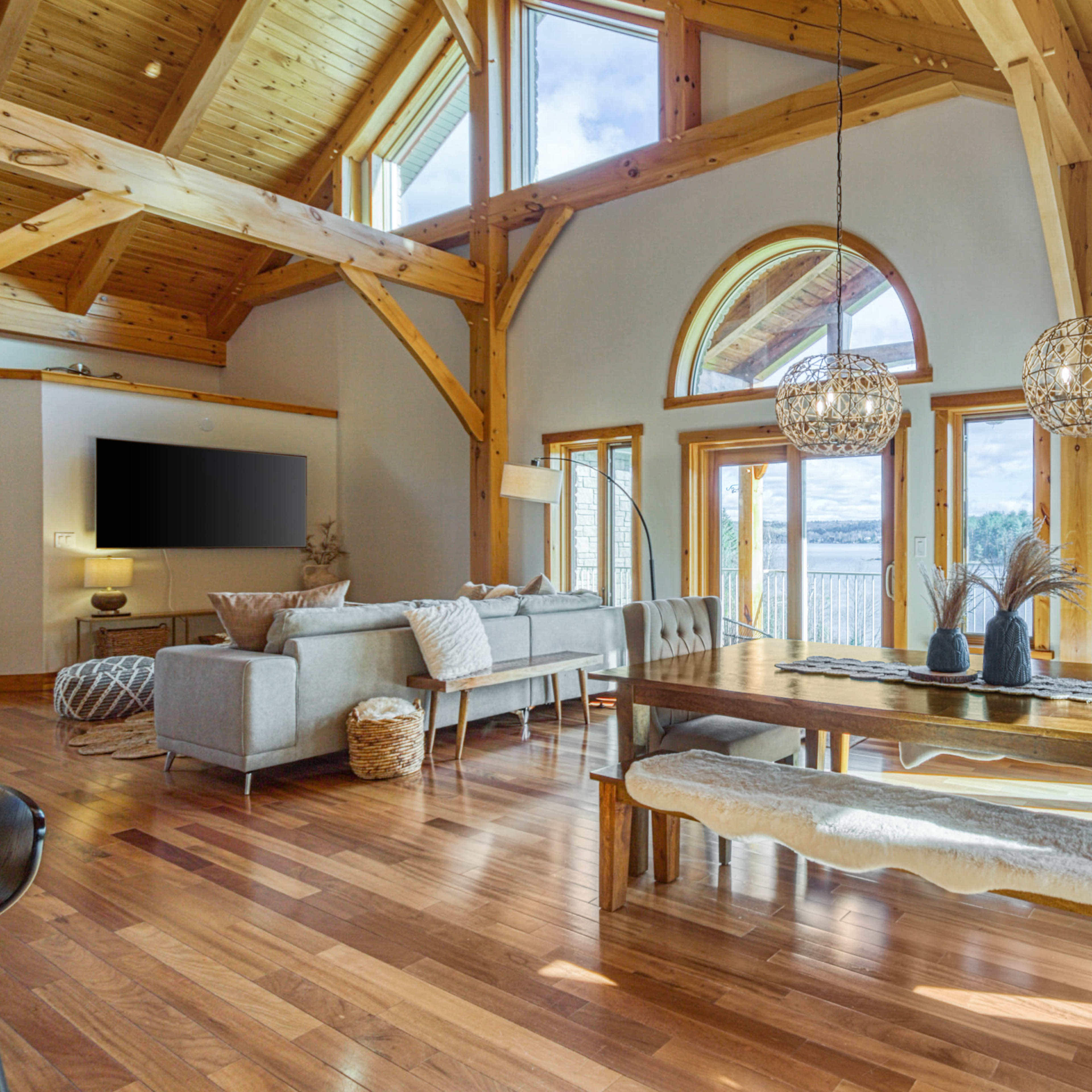 A bright, spacious interior features wooden beams, large windows overlooking a body of water, and a dining table surrounded by comfortable seating.