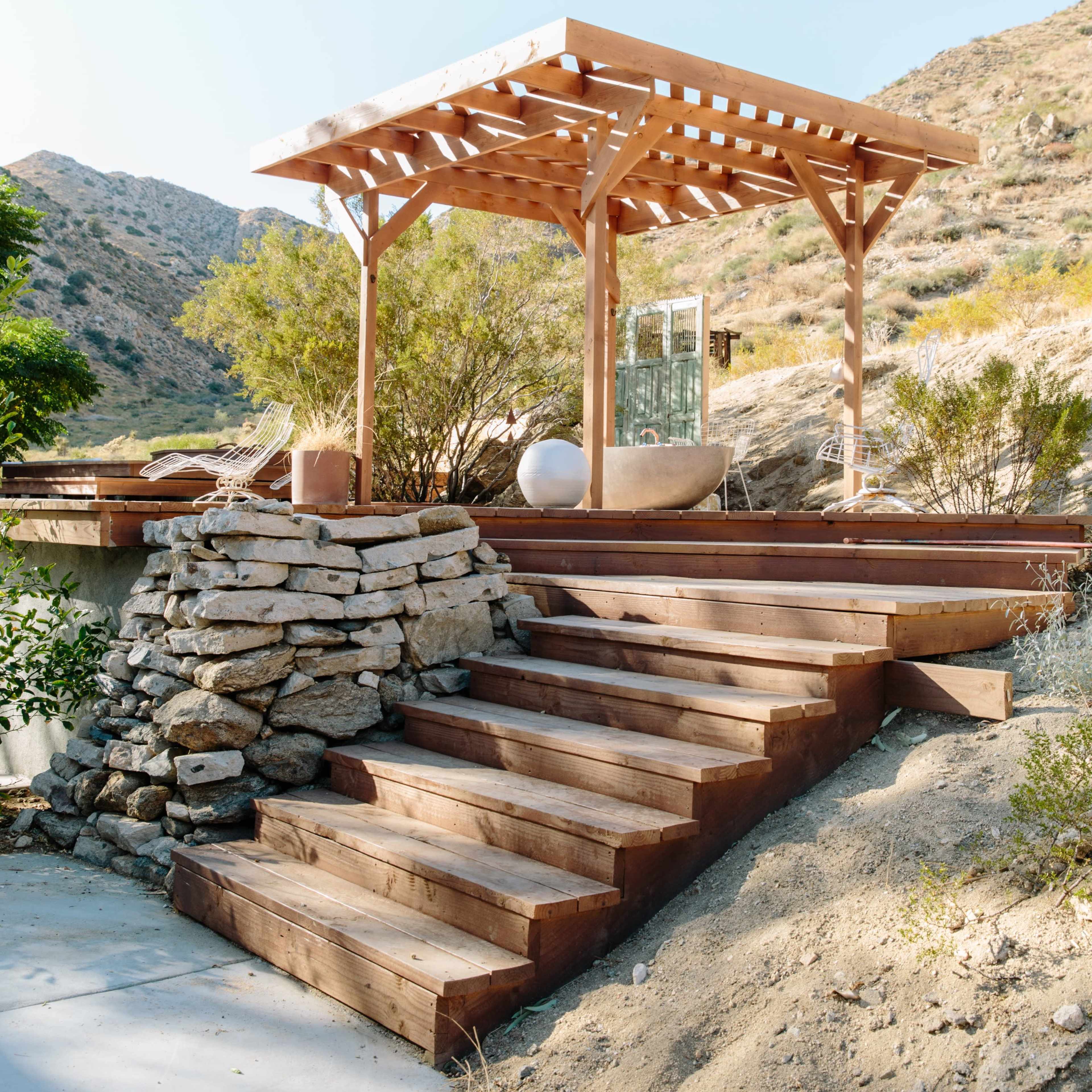A wooden pergola sits atop a landscaped hillside, surrounded by rocky steps and native vegetation.