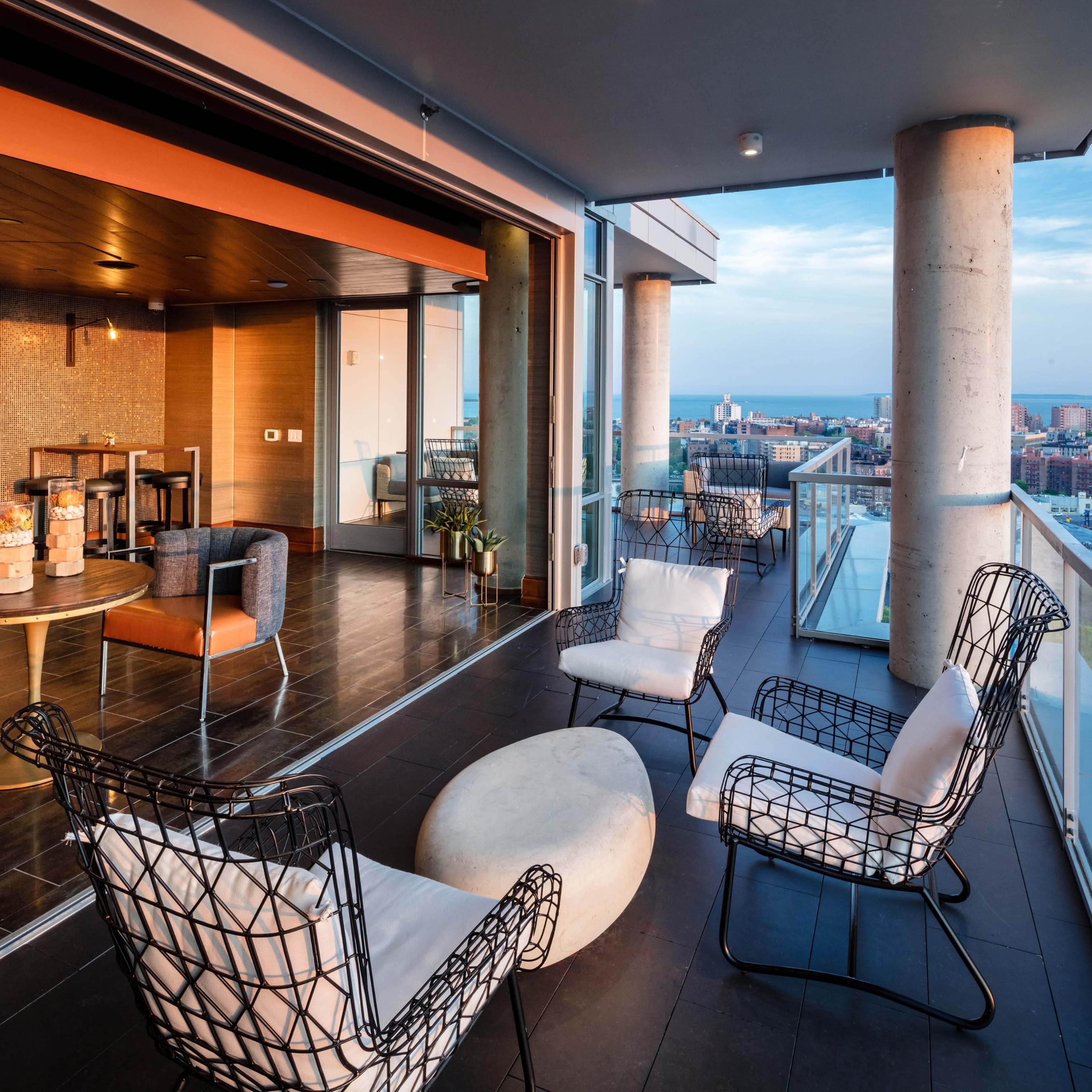 A modern balcony features wire chairs, a circular table, and a view of the city skyline and train tracks.