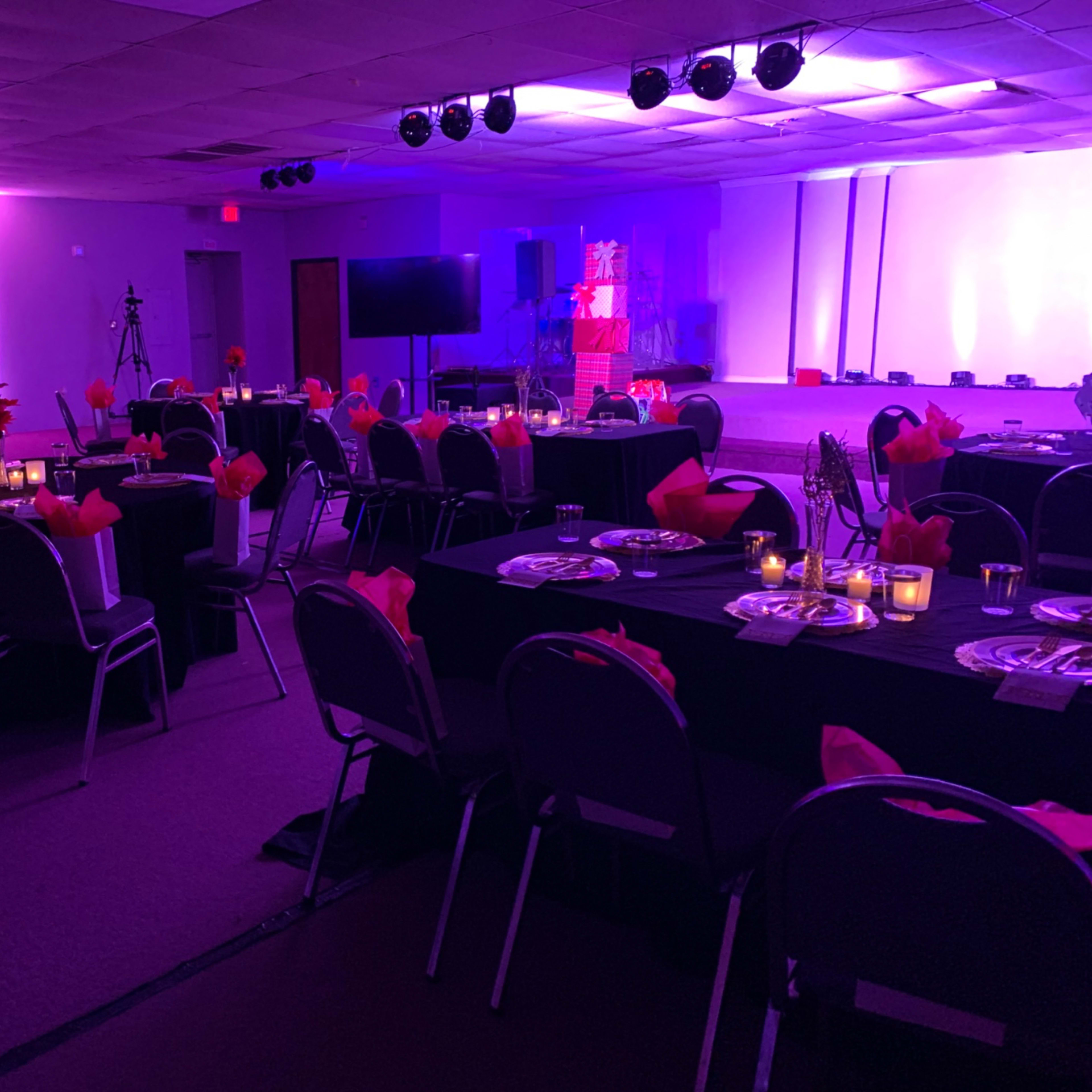 A banquet hall set up with tables covered in black cloth, adorned with pink napkins and lit candles, all under purple accent lighting.