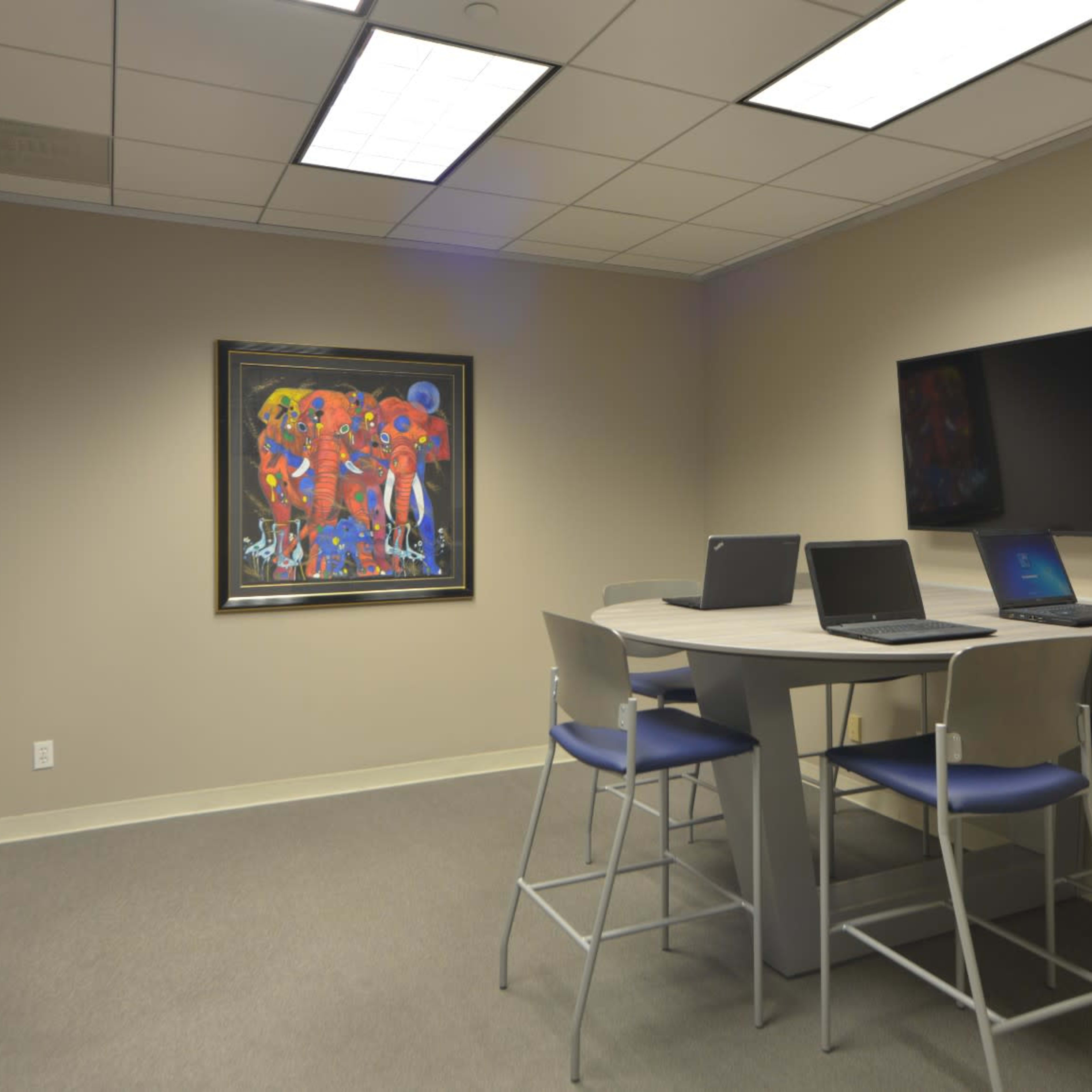 A conference room with two high tables and chairs, featuring three laptops, a mounted television, and a vibrant piece of artwork on the wall.