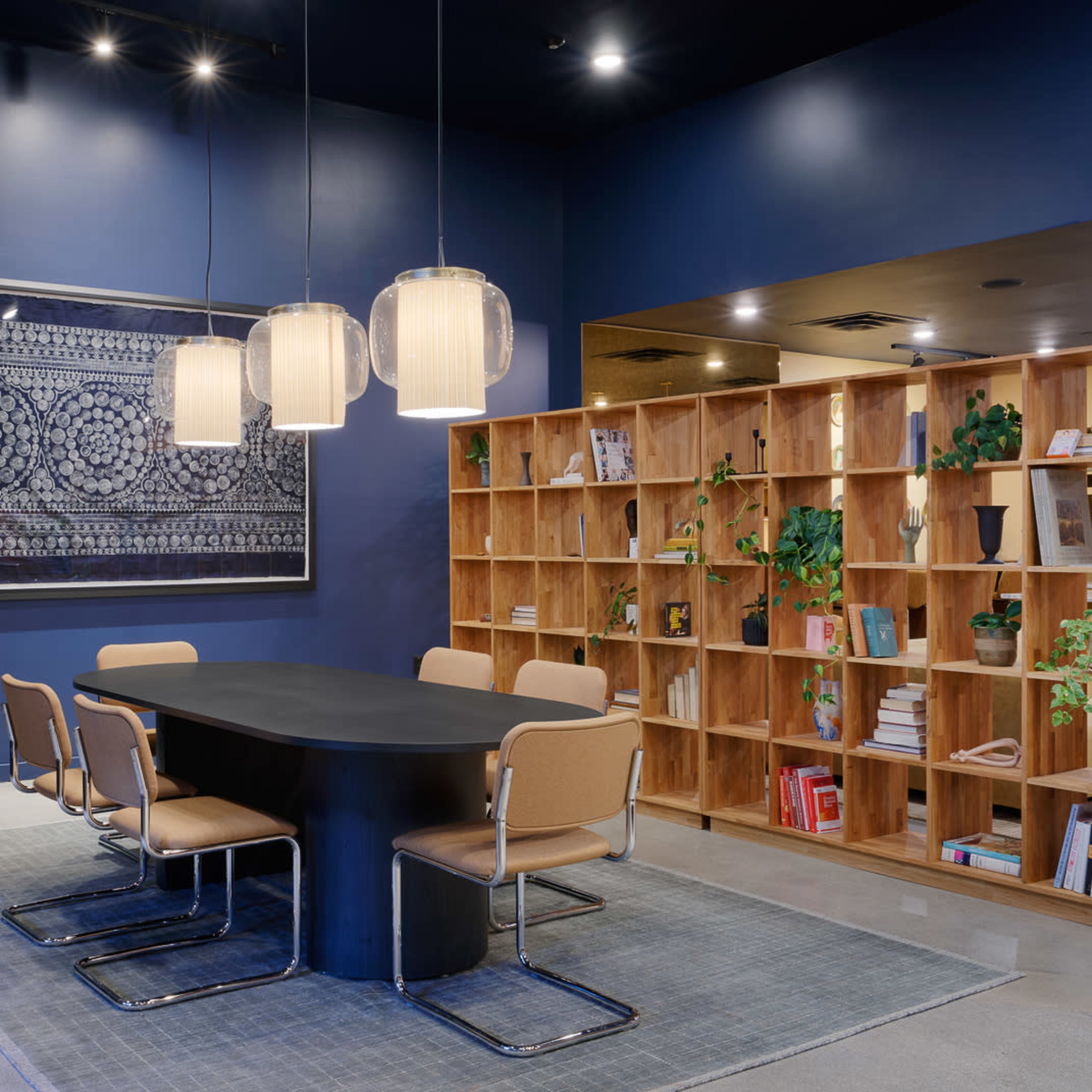 A modern conference room features a large oval table surrounded by beige chairs, with a wooden bookshelf filled with books and plants against a dark blue wall.