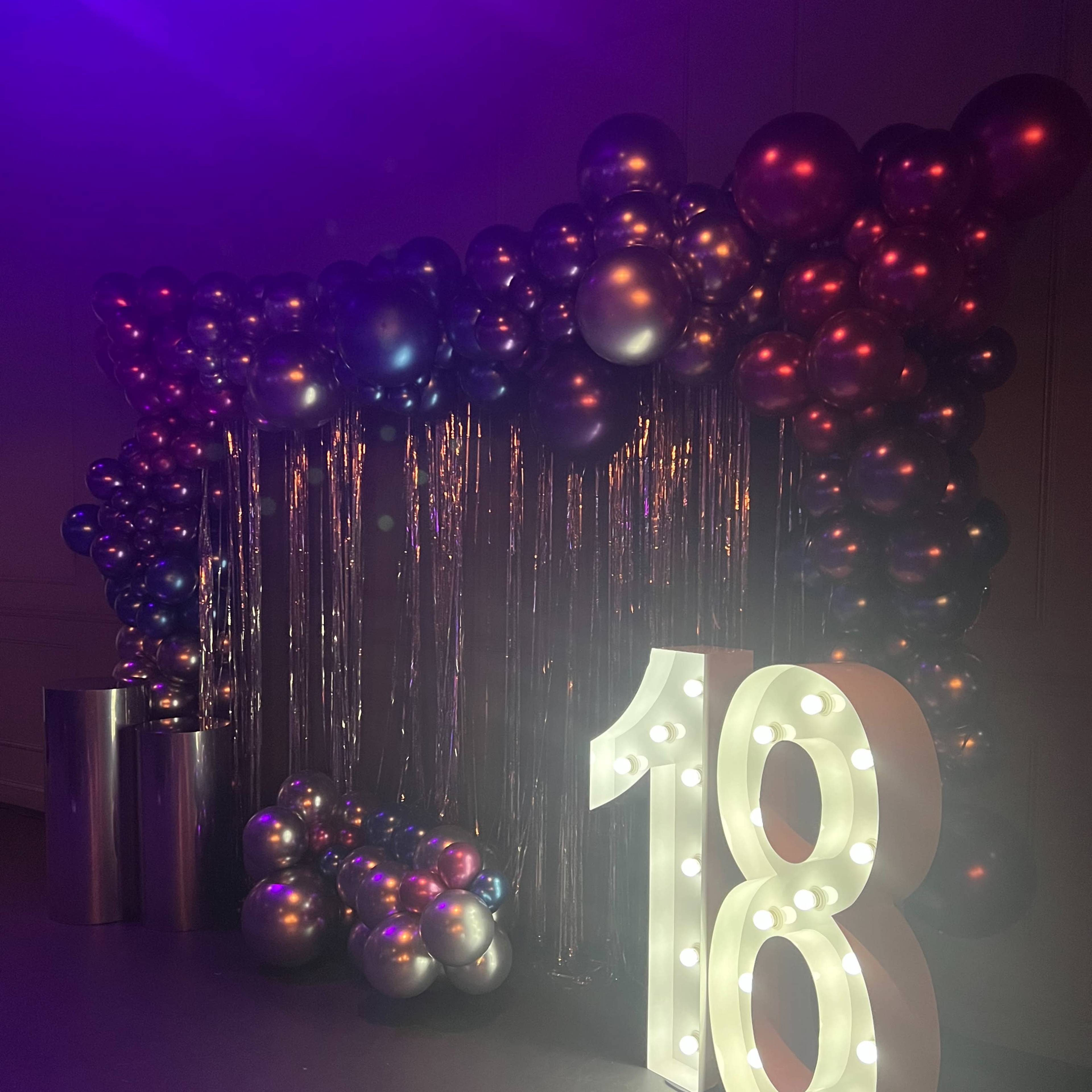 A decorative setup for an event, with a large illuminated "18" sign surrounded by a backdrop of metallic balloons and a curtain of shiny fringe.