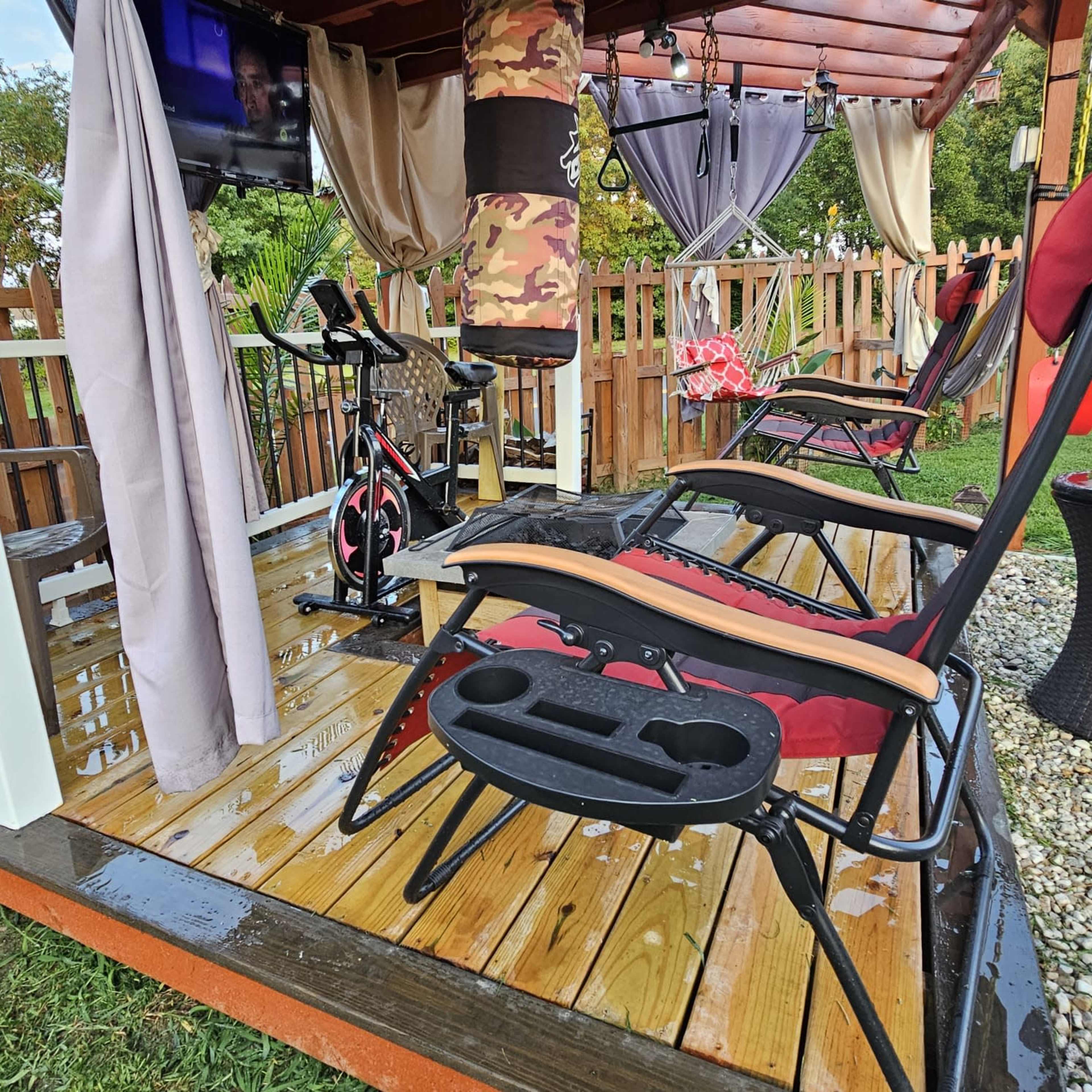A sheltered outdoor space features reclining chairs and an indoor exercise bike on a wooden deck, with a television mounted nearby.