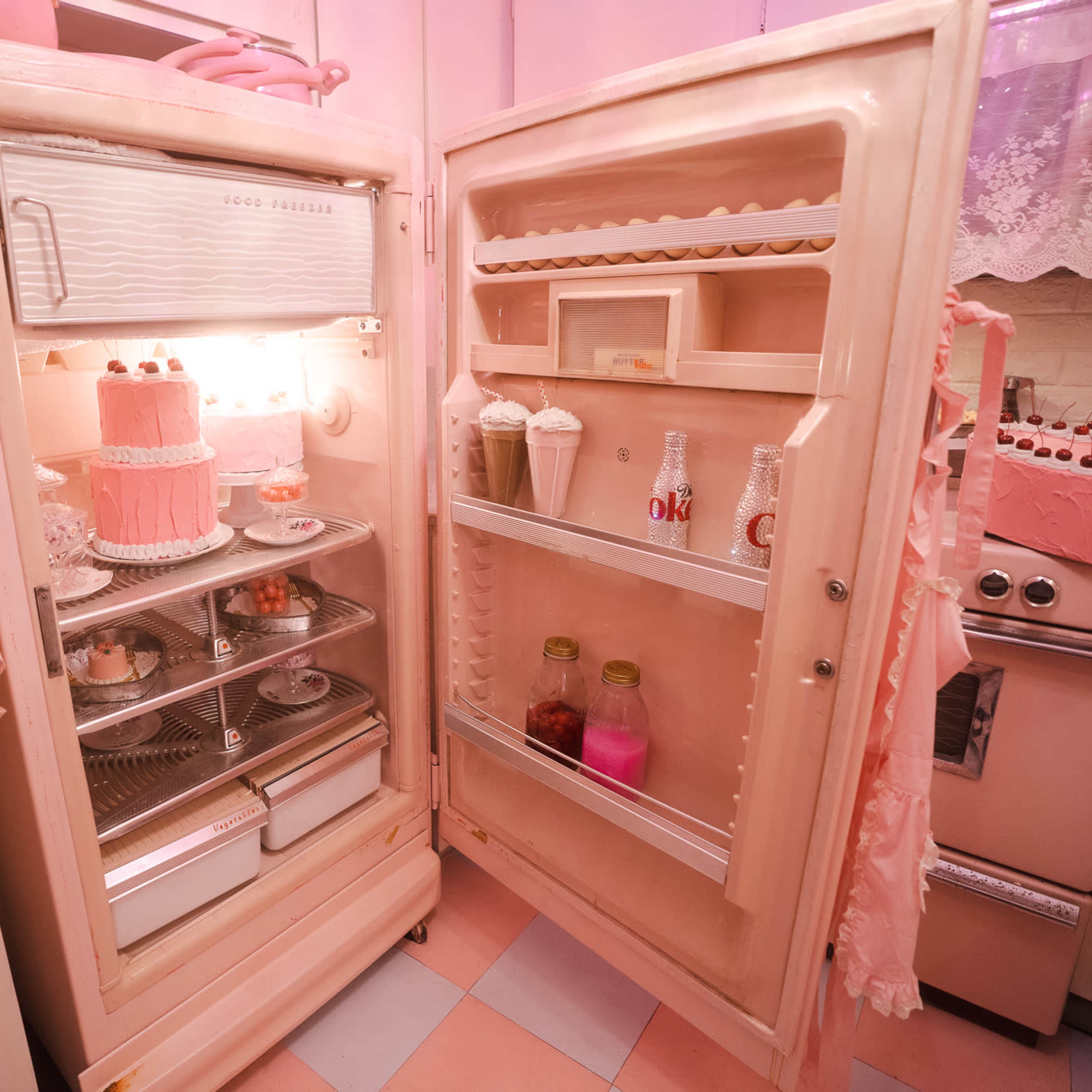 A vintage pink refrigerator with its doors open, revealing a cake, drinks, and desserts inside, set in a pink-themed kitchen.